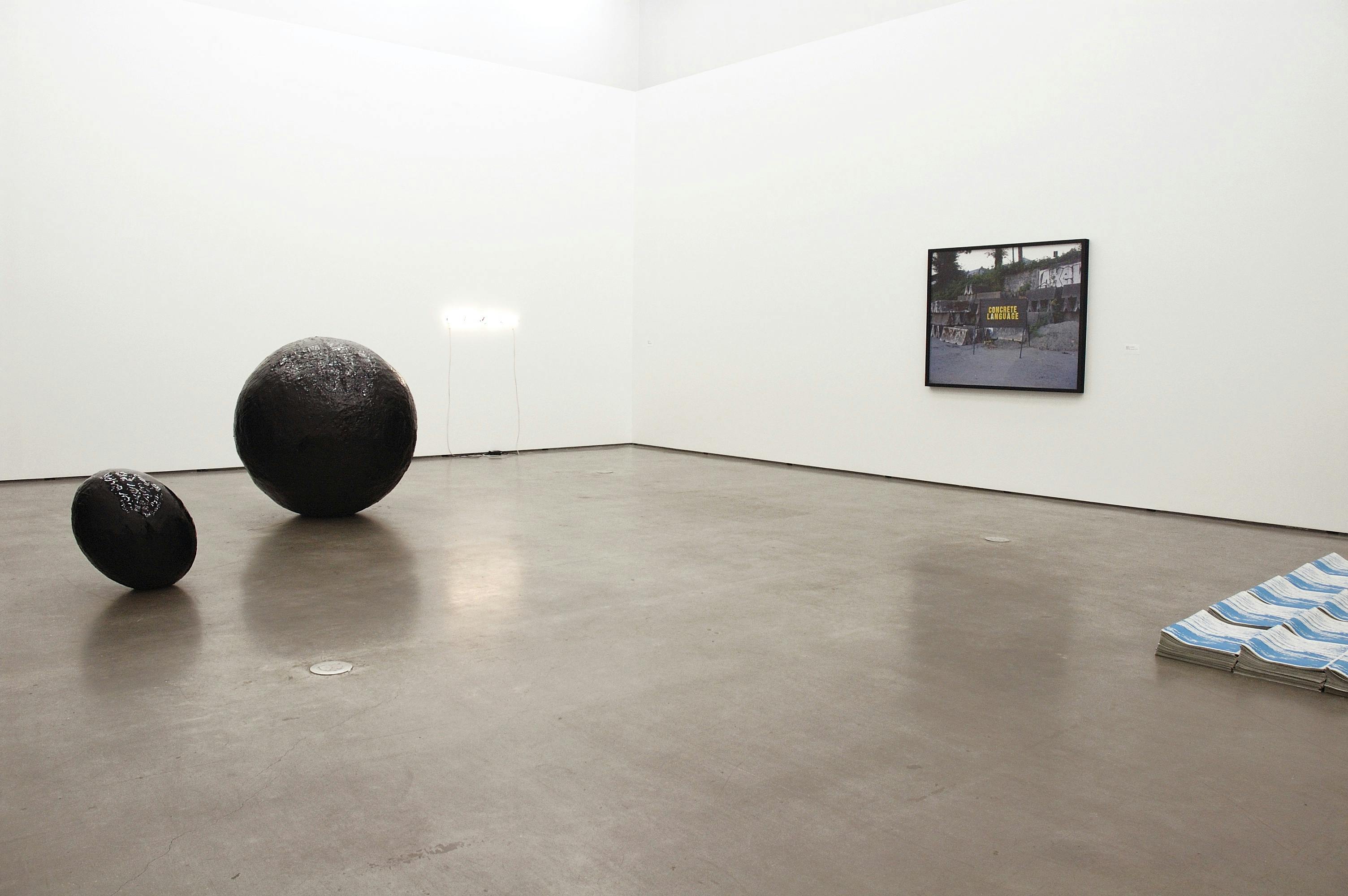 This is an installation shot of a group exhibition titled Concrete Language. A large-sized photograph is mounted on a wall. On the floor, two round-shaped black sculptures are placed. 