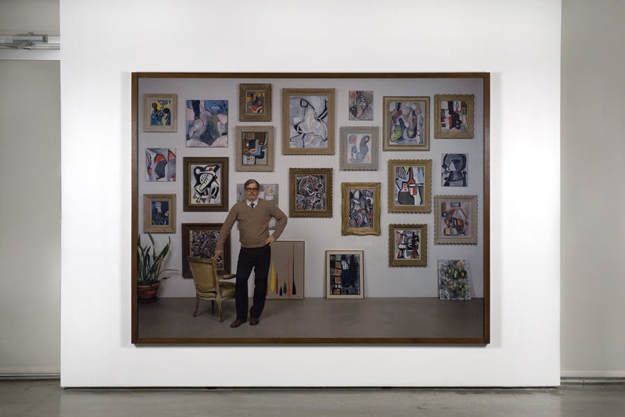 A large scale framed photograph hangs on a gallery wall. The image depicts Rodney Graham posing as a painter in front of a wall of abstract paintings hung in salon style. He is leaning by a chair.