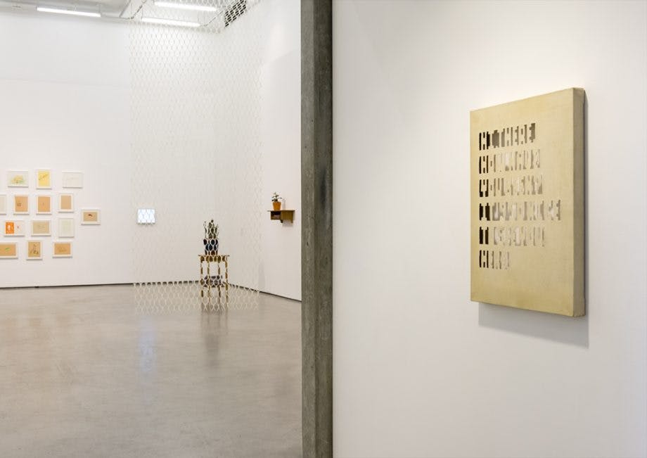 Various artworks installed in a gallery from framed drawings to two sculptures. A stretched canvas on the right has text cut into the surface.