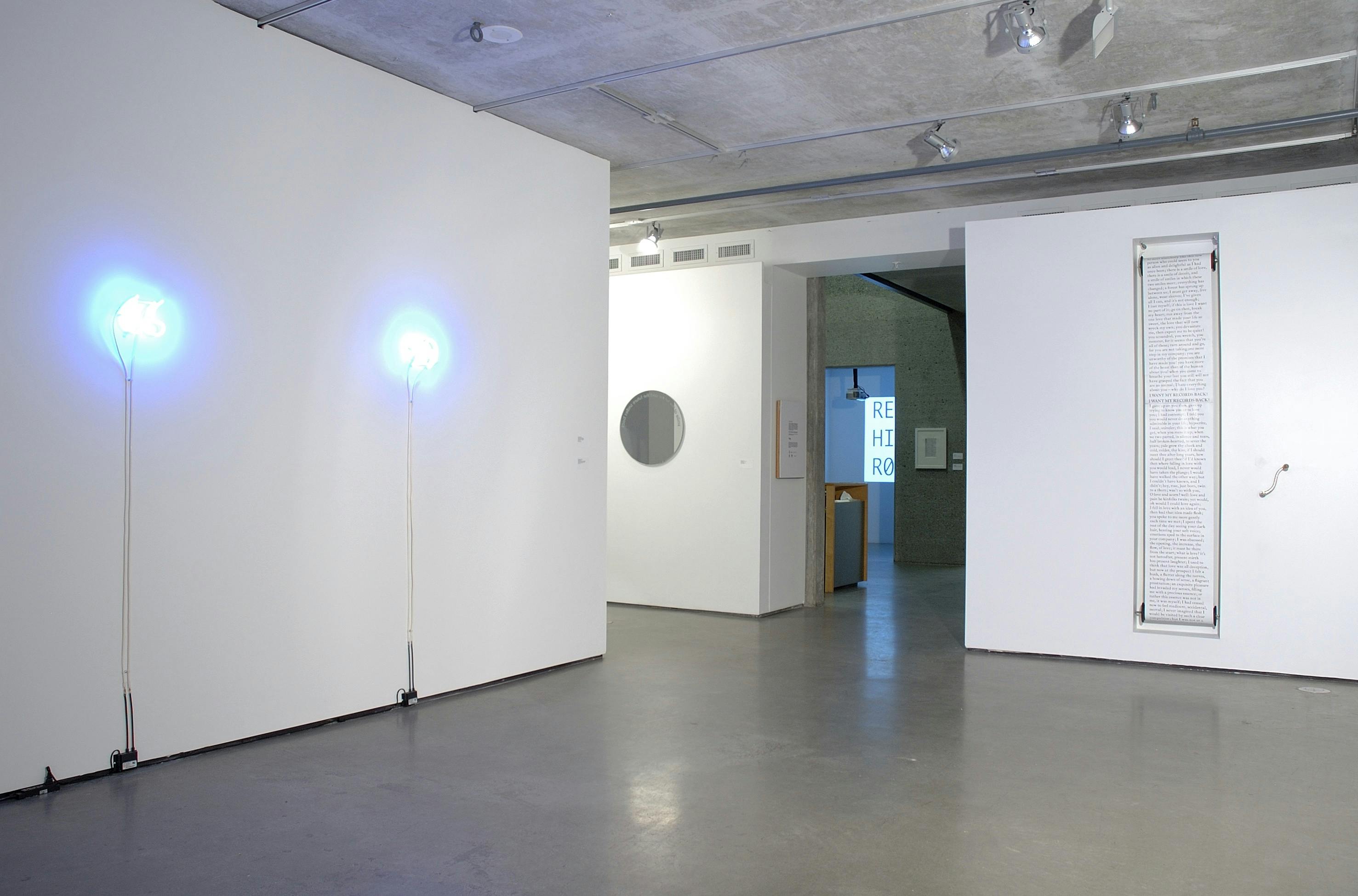 This is an installation shot of a group exhibition titled Concrete Language. A tall and narrow white paper is mounted on the wall beside the gallery entrance. A neon sign is placed on the other wall. 