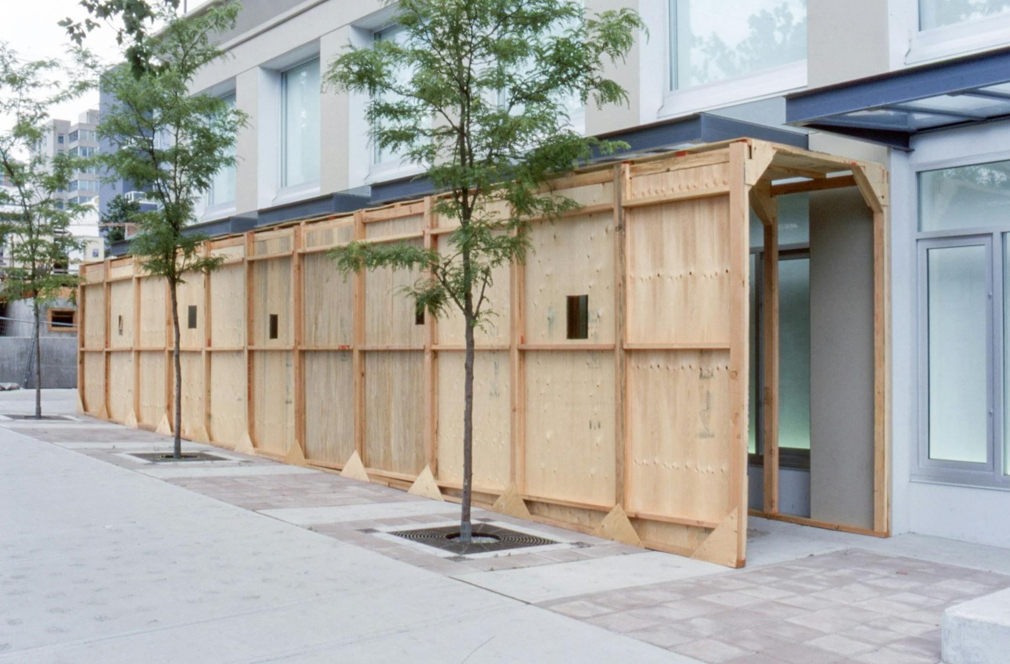 This is Brian Jungen’s installation resembling construction hoardings. Wood tiles with some small windows cover the tunnel. Some trees are planted in front of this installation.
