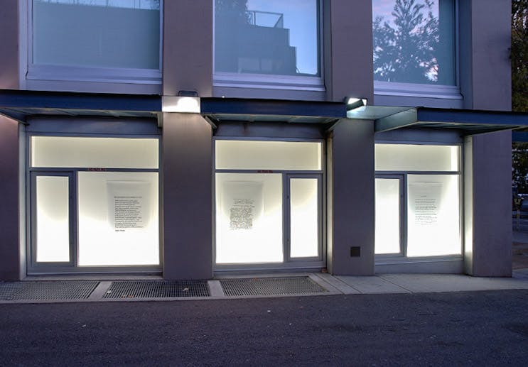 An installation image of a work by Daniel Olson in CAG’s window spaces facing a sidewalk. Three large pieces of white paper, on which some typed texts are visible, are mounted on an illuminated wall behind glass windows.