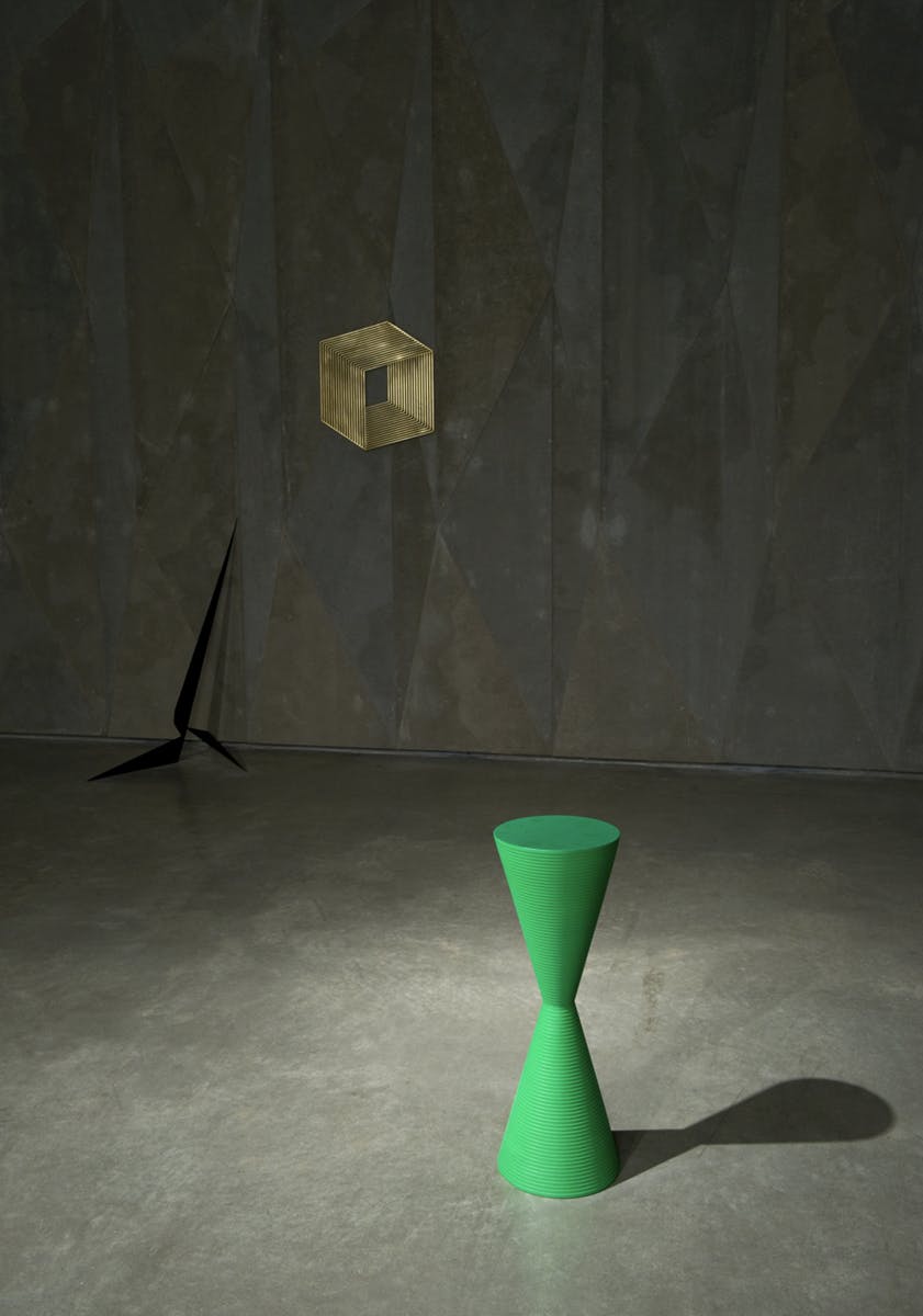 Two sculptures and one drawing are visible in this image. An hourglass-shaped green sculpture and a thin, black propeller-shaped sculpture sit on the floor below a golden cube drawn on the wall.