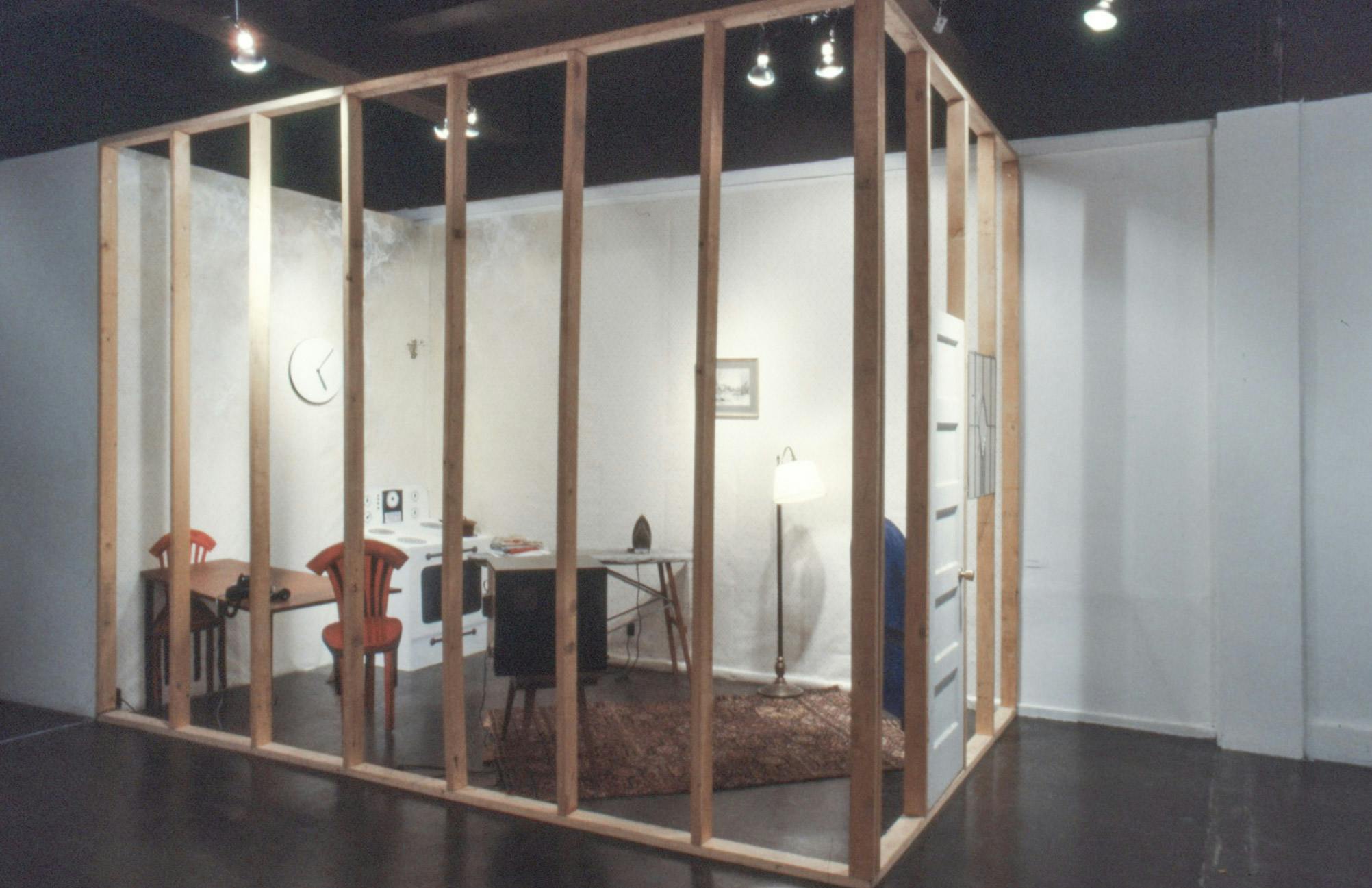 A closeup of an installation in a gallery. The installation resembles a living space with sculptures of various items including a TV, a stove, chairs, an ironing board, a rug, and a lamp.