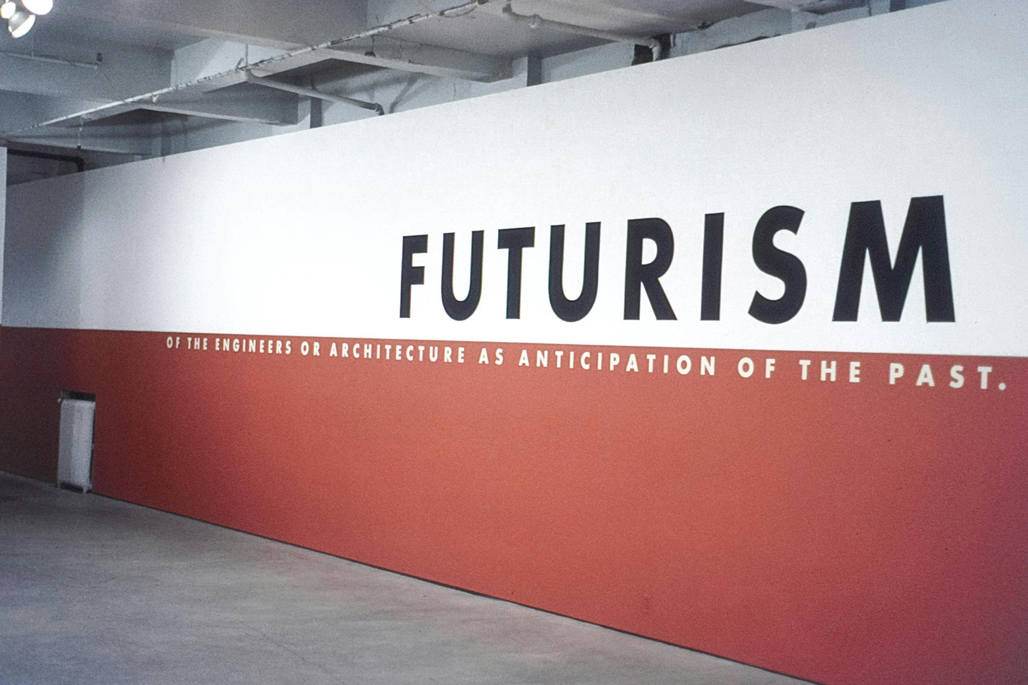 A wall of a gallery with black and white text. The top half of the wall is white and the bottom is red. The text reads "Futurism: of the engineers or architecture as anticipation of the past." 
