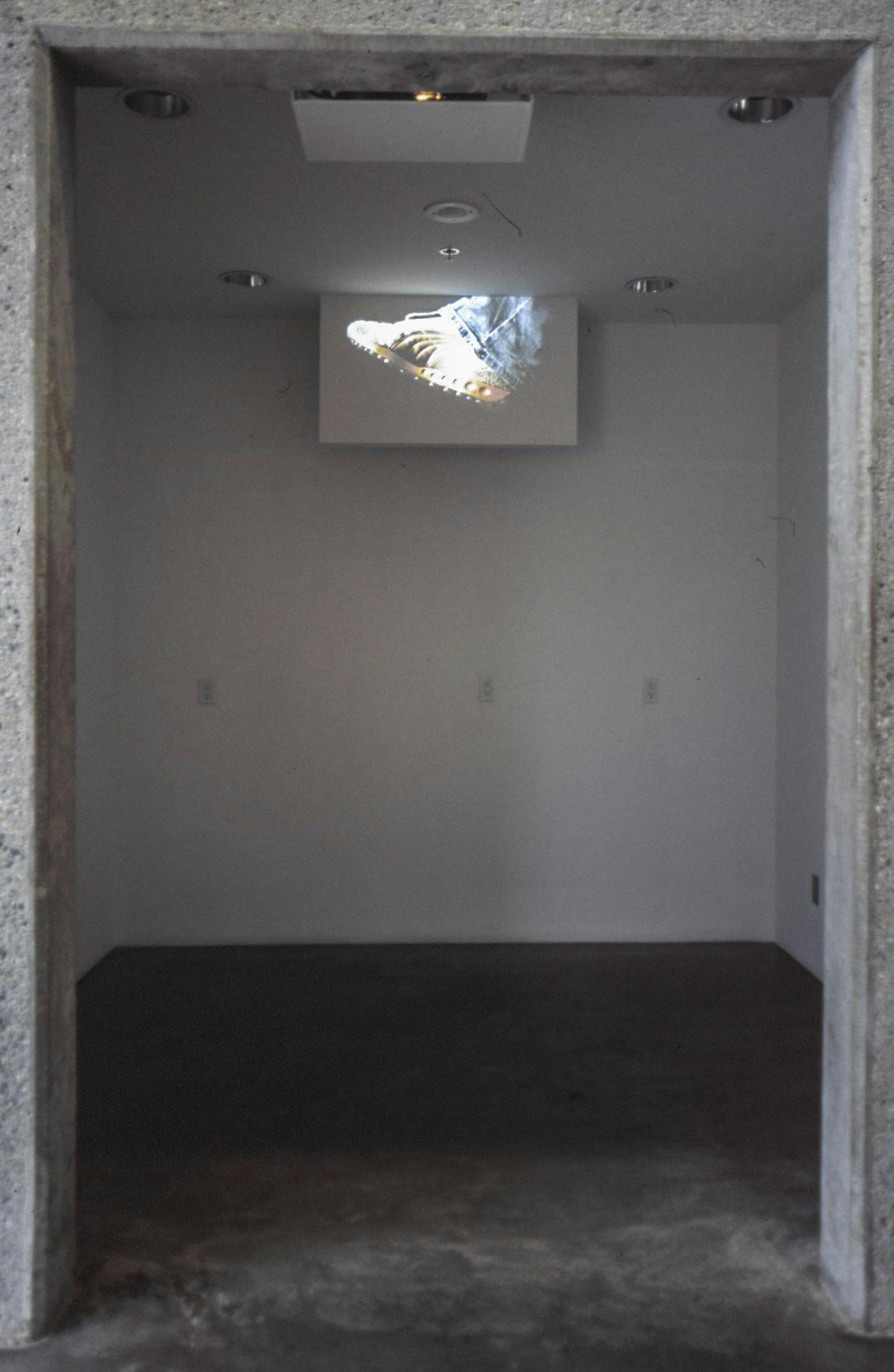 A single-channel video of a runner’s foot is projected at the top of the wall in a gallery space. The video shows a foot wearing a white sneaker and blue jeans running in the dark.