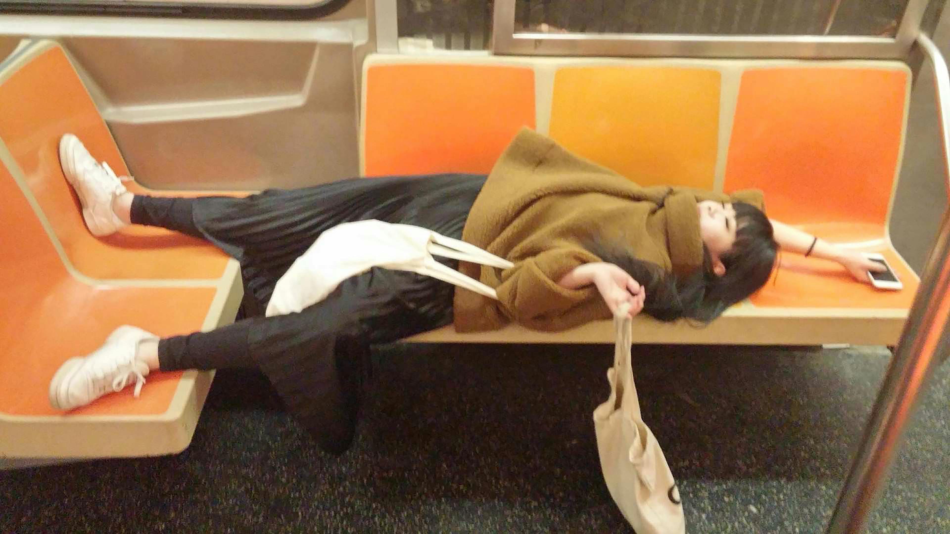 A portrait of Qian Cheng lying across the orange seats of a subway car. Qian is wearing a brown turtleneck sweater that is covering her chin. One of her arms is stretched behind her head holding an iPhone; the other is holding two tote bags.