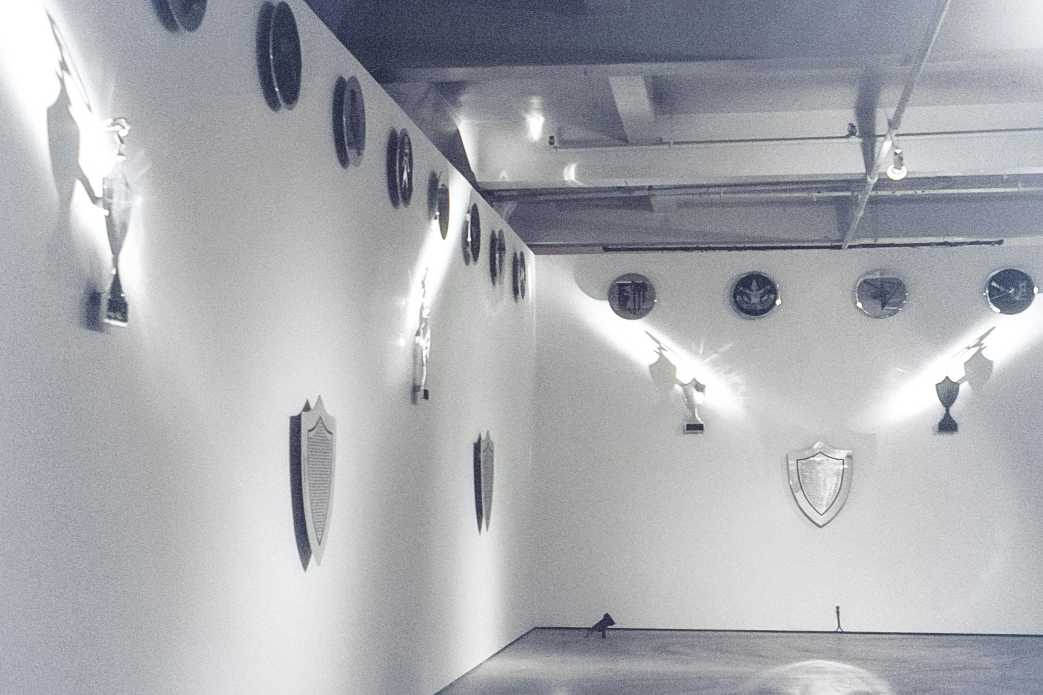 The corner of a gallery space with metal artworks resembling family crests and shields. The works are all silver with black details and the light in the room reflects dramatically from their surface.