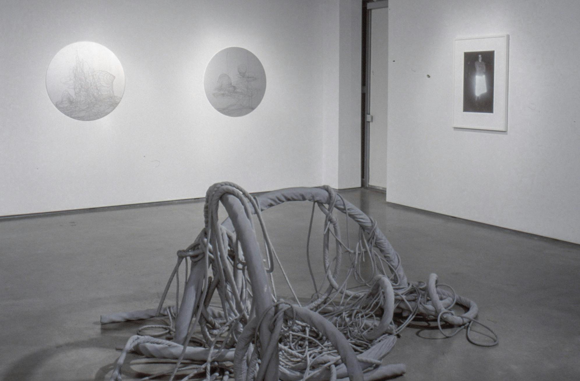 Four artworks are installed in a gallery space. A large grey sculpture sits on the floor. A  framed black and white photograph and two circular shaped sculptures are mounted on the walls.