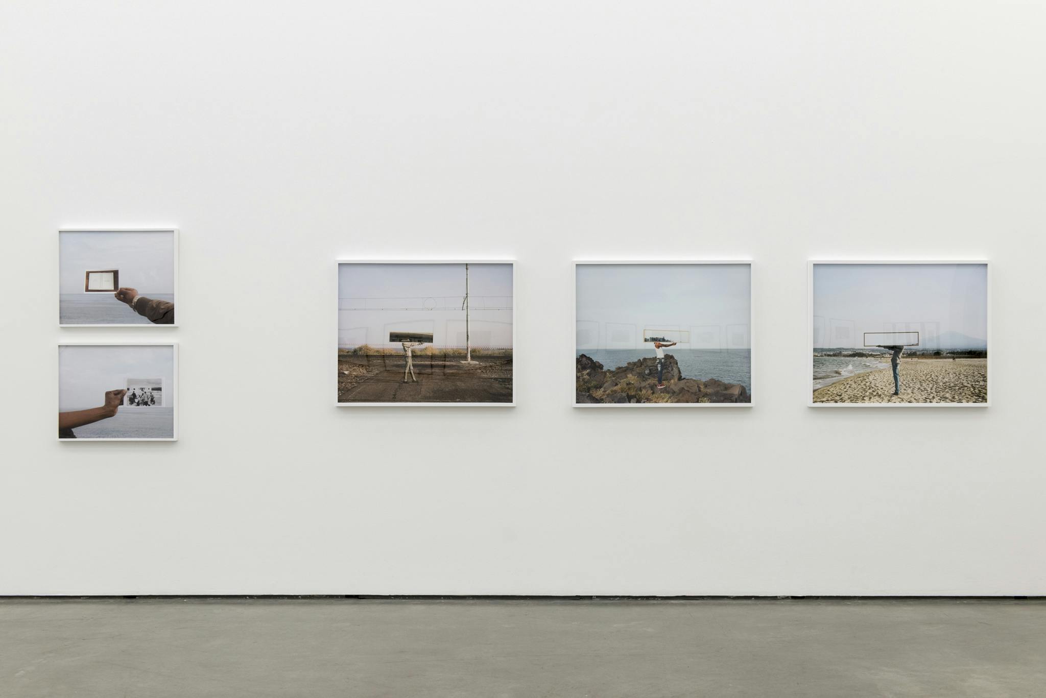 Five framed photographs hang on the gallery wall. Three are of a person holding a mirror in front of their head, outdoors. Two photographs are of a hand holding another photo in front of the ocean. 