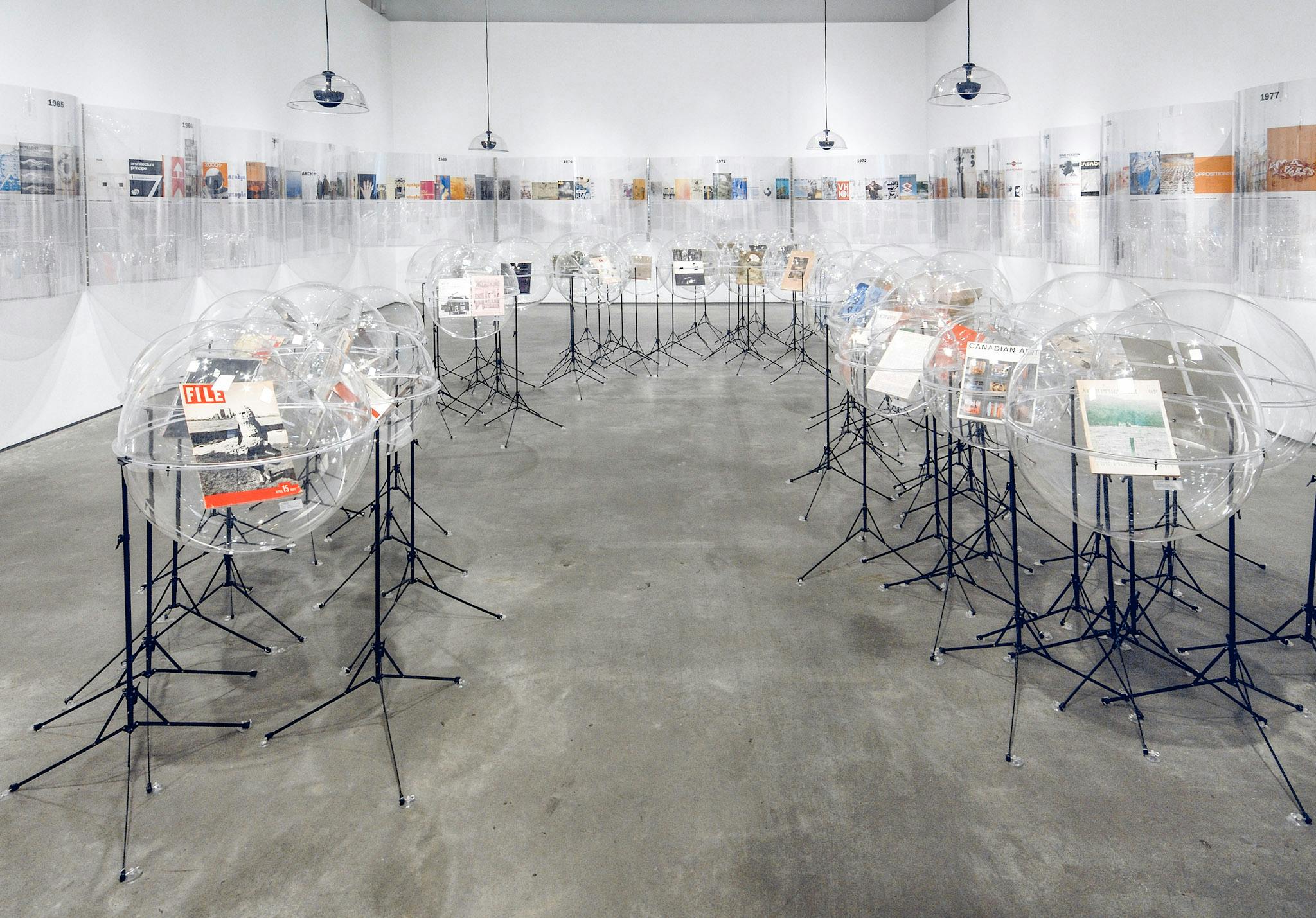 Many sculptures are installed in a gallery. They are magazines placed inside transparent rounder-shaped plastic cases. Those cases are supported by black metal legs similar to those of music stands. 