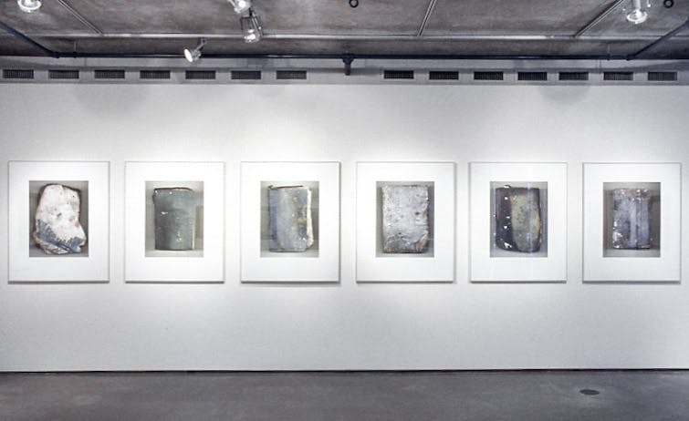 Six large photos in silver frames, on a white wall. The photos show different burnt and deteriorated books. The photos are mostly shades of white, black, and green. 