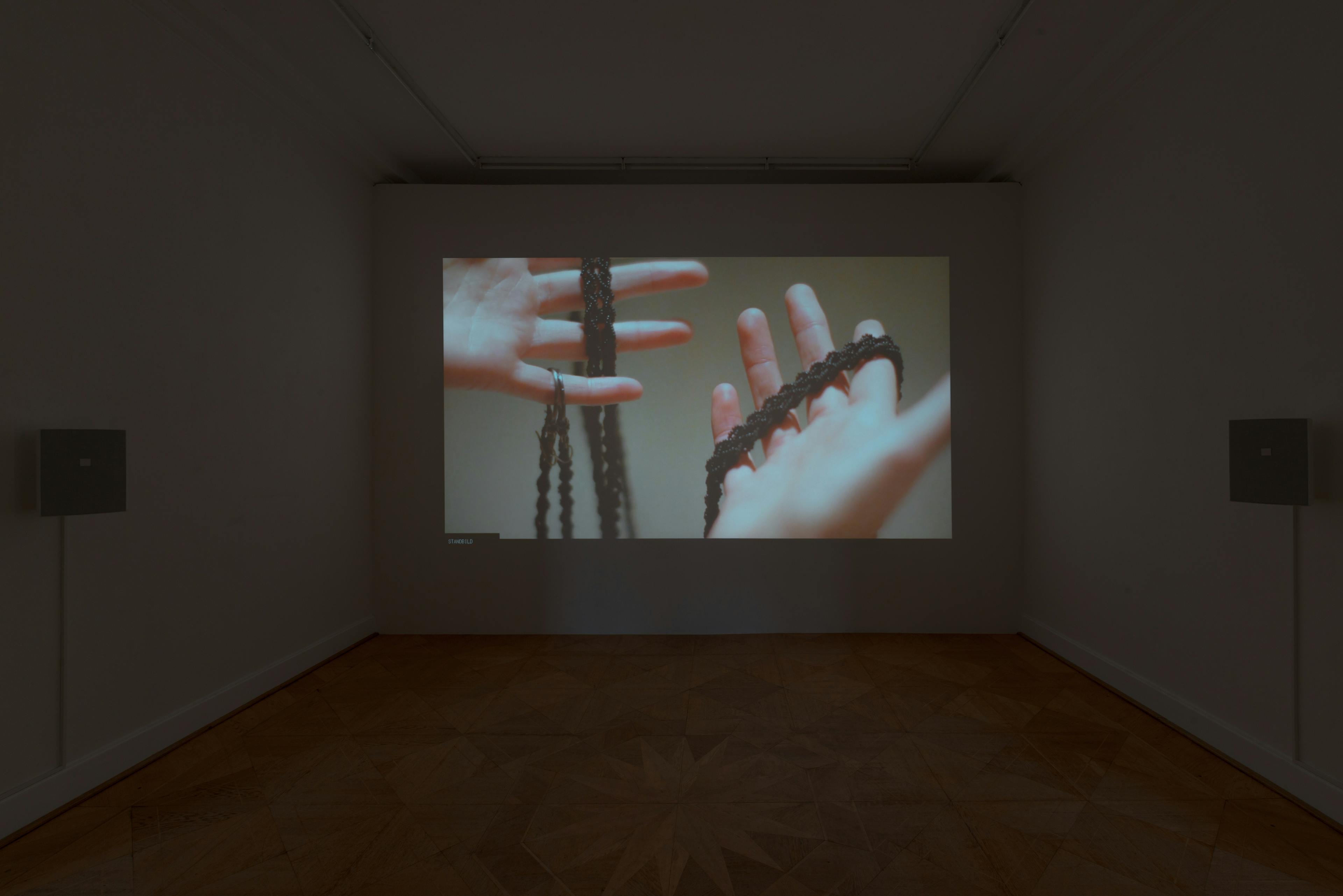 In a darkened gallery space, a single-channel video work is projected on a white wall at the end of the room. The video shows a pair of hands holding a beaded rope.