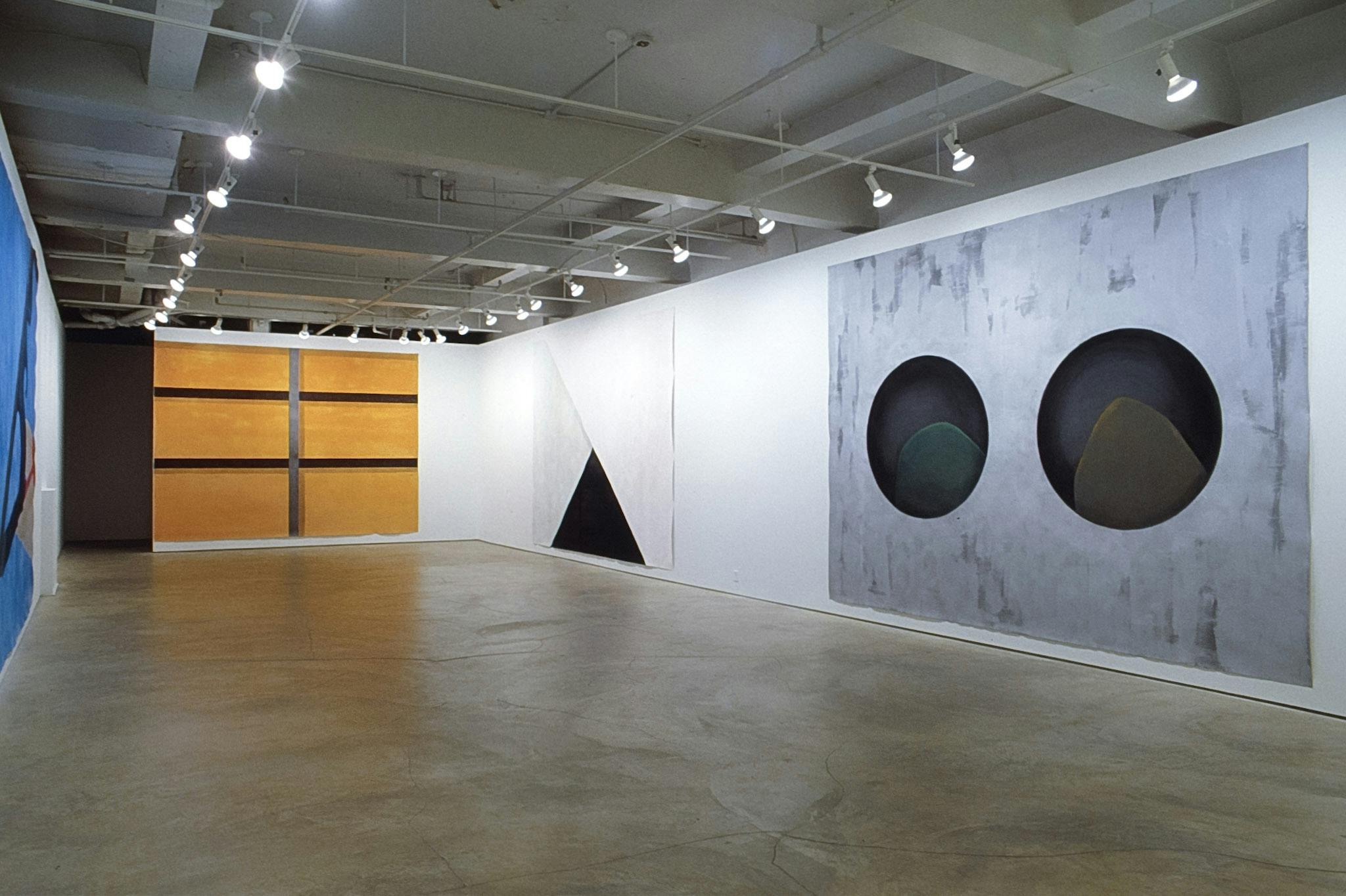 Four paintings are installed in the gallery. Two on the right side wall depict black geometric shapes. The one on the inner wall shows six orange rectangles. The one on the left side wall is blue.