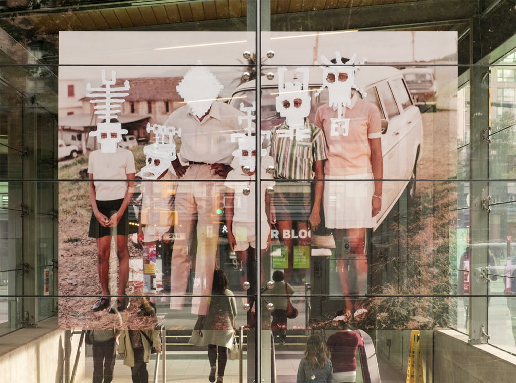 A large-scale photograph in vinyl installed on the glass exterior of Yaletown-Roundhouse Station. In the image, a family stands in front of a car. Mask-like shapes have been painted over their faces.