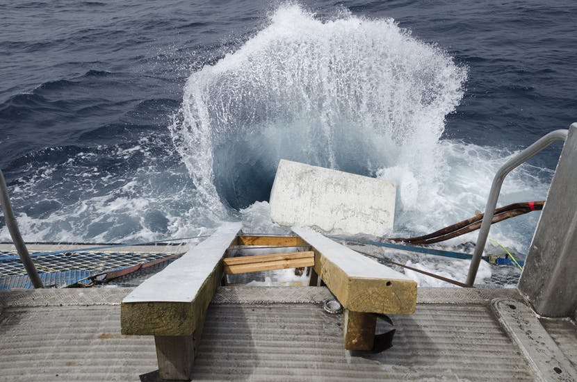 A photograph of a rectangular stone falls into a dark blue ocean creating a large splash. An industrial metal structure fills the foreground of the photograph. 