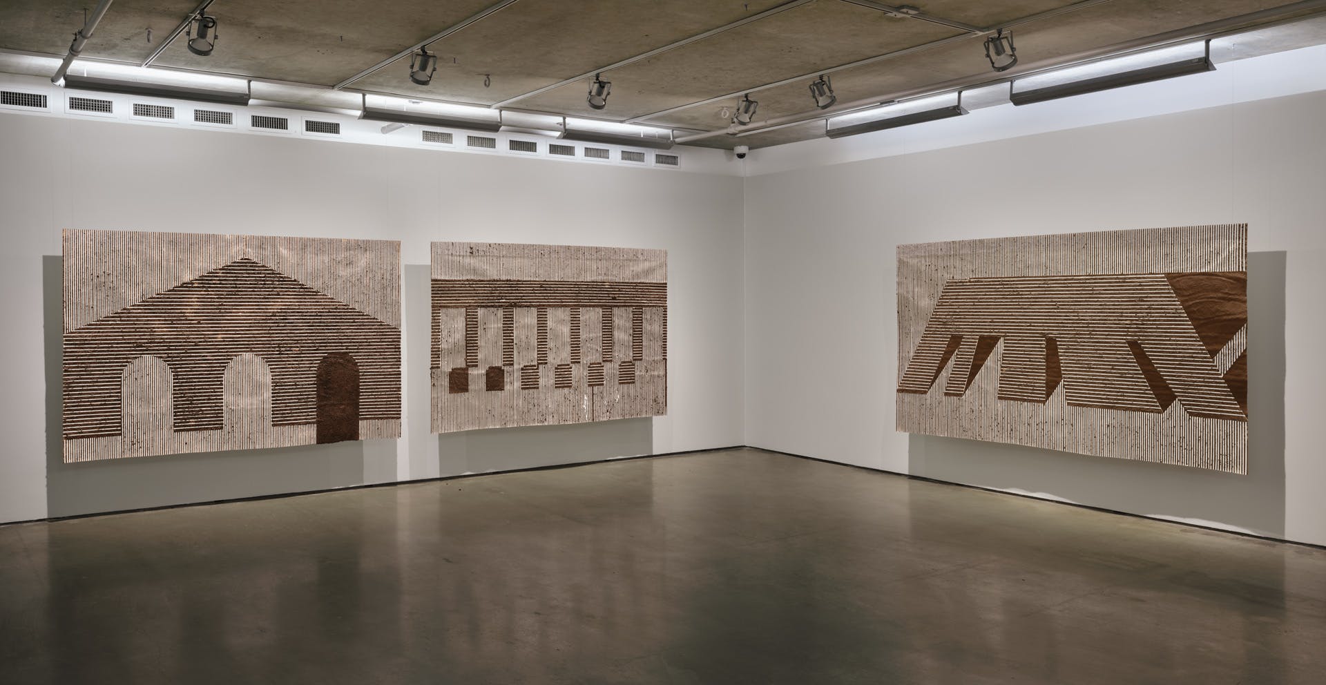Three large scale drawings are suspended against the two sides of the corner wall of a gallery space. The drawings of architectural forms are made with striations of brown adobe mud on white paper. 