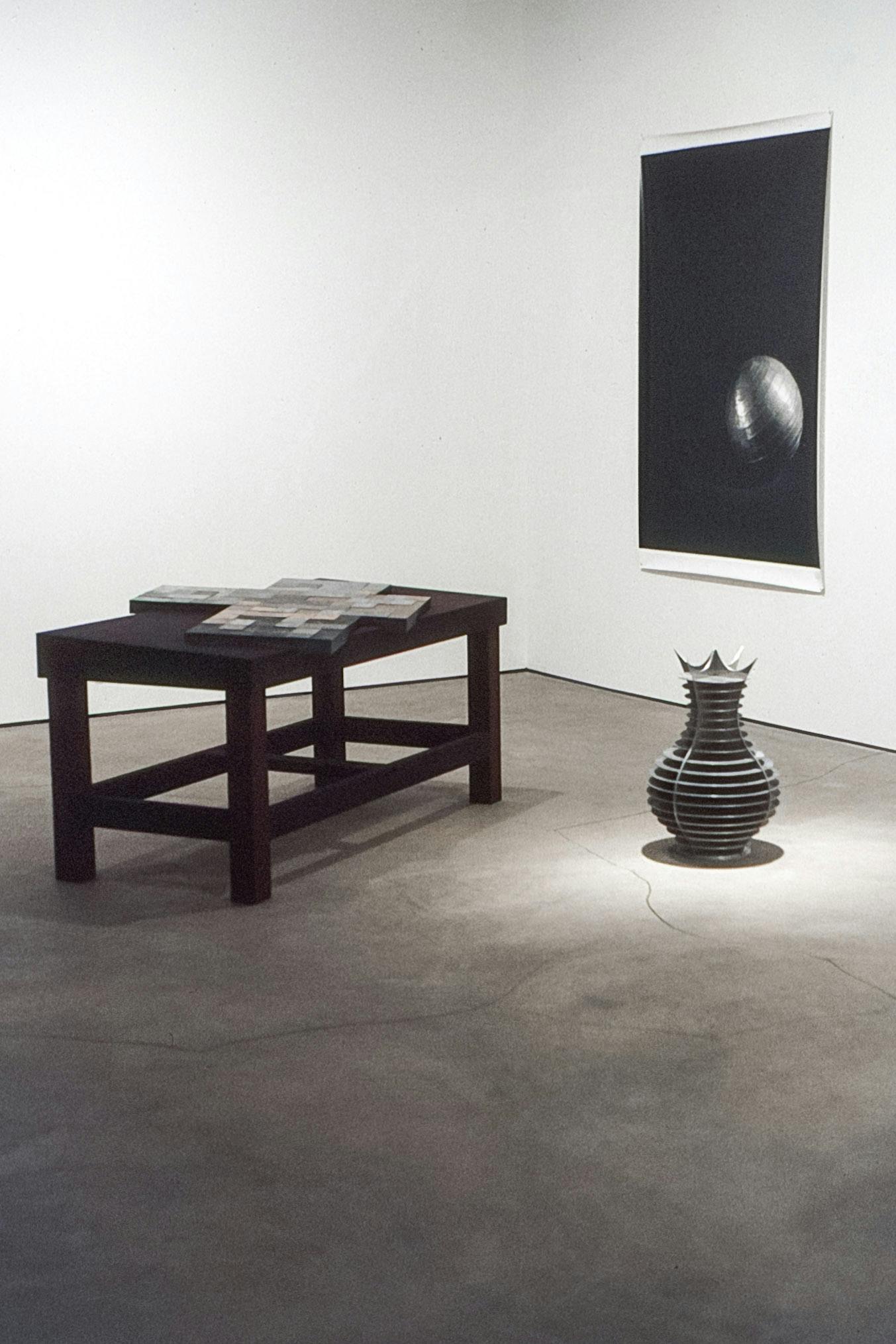 The corner of a gallery space. A large photo of a silver ball in a dark room is on a wall. On the floor, a table with an asymmetrical cross painting on it, and a vase with metal detailing beside it.