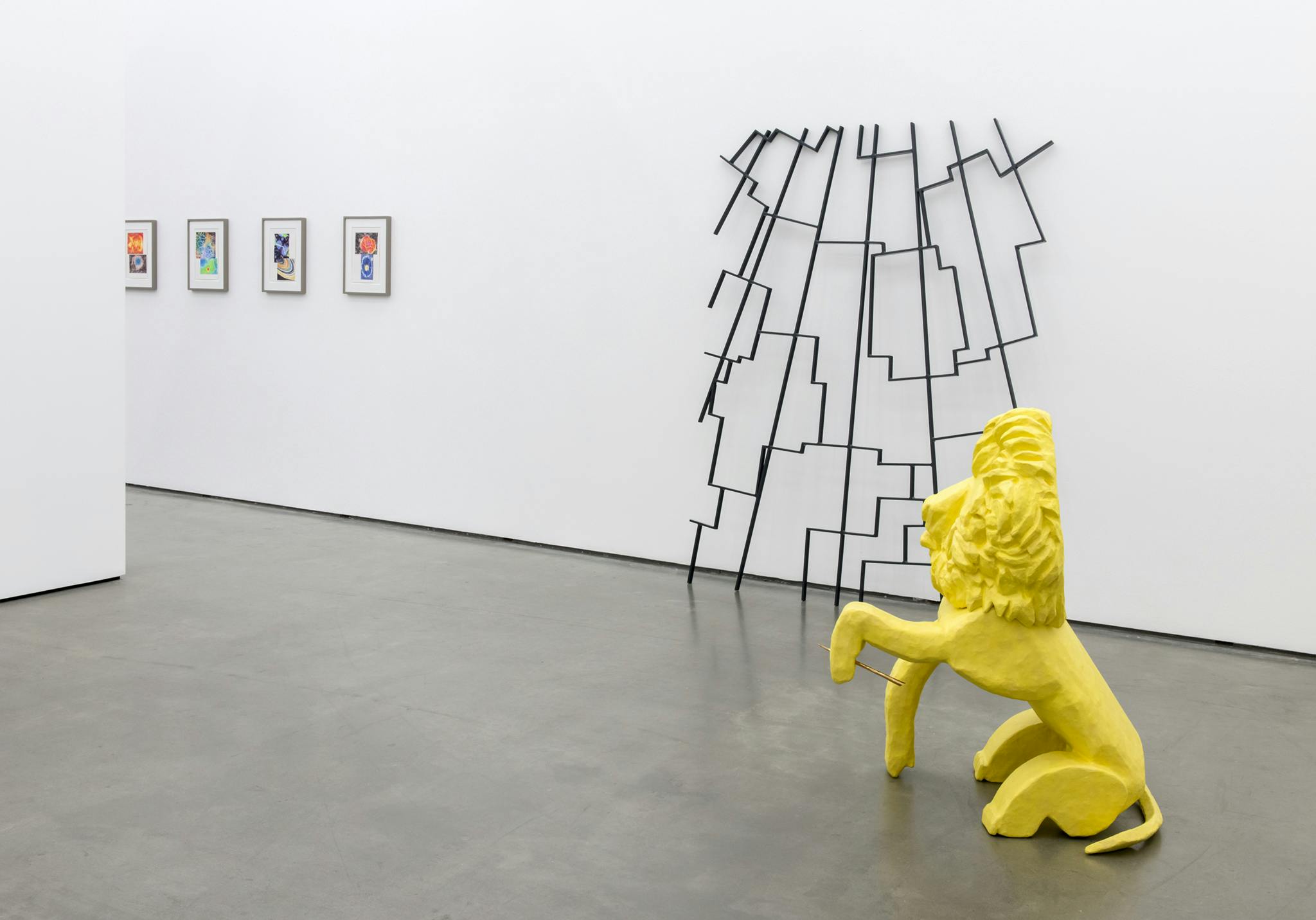 A yellow lion sculpture sits on a gallery floor. Behind it, a thin, black steel sculpture leans against a wall. Several framed, small-scale colour drawings are installed on the same gallery wall. 
