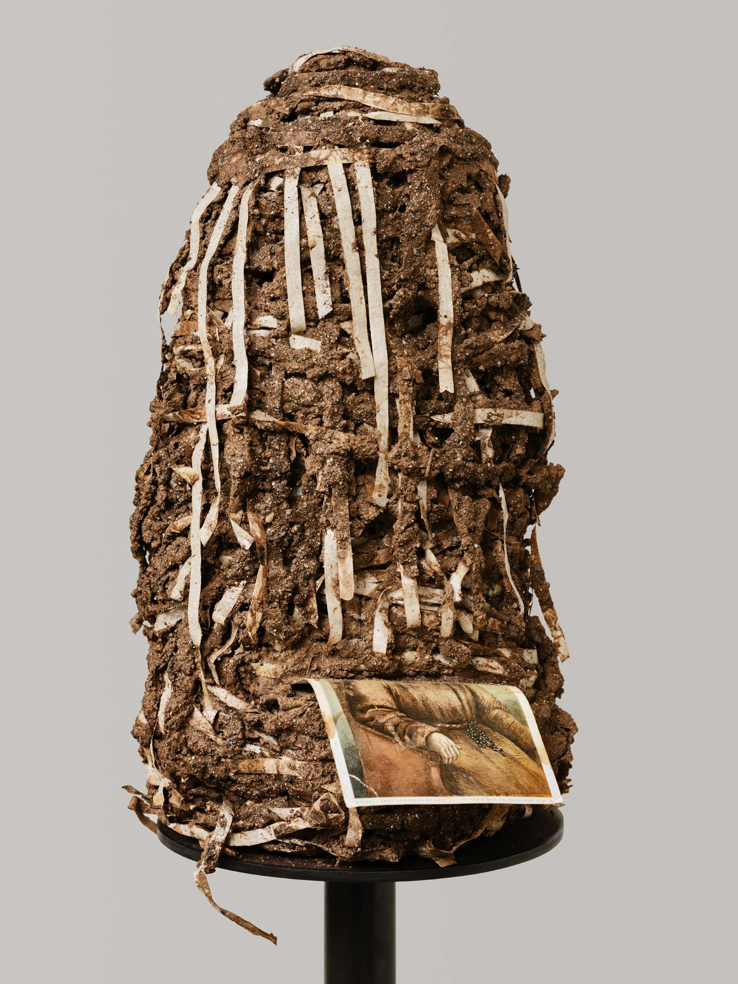 A mound-like sculpture made from layers of adobe mud and strips of tape. A postcard with an image of a figure dressed in a religious habit is wedged into the mound, leaving only the torso visible.