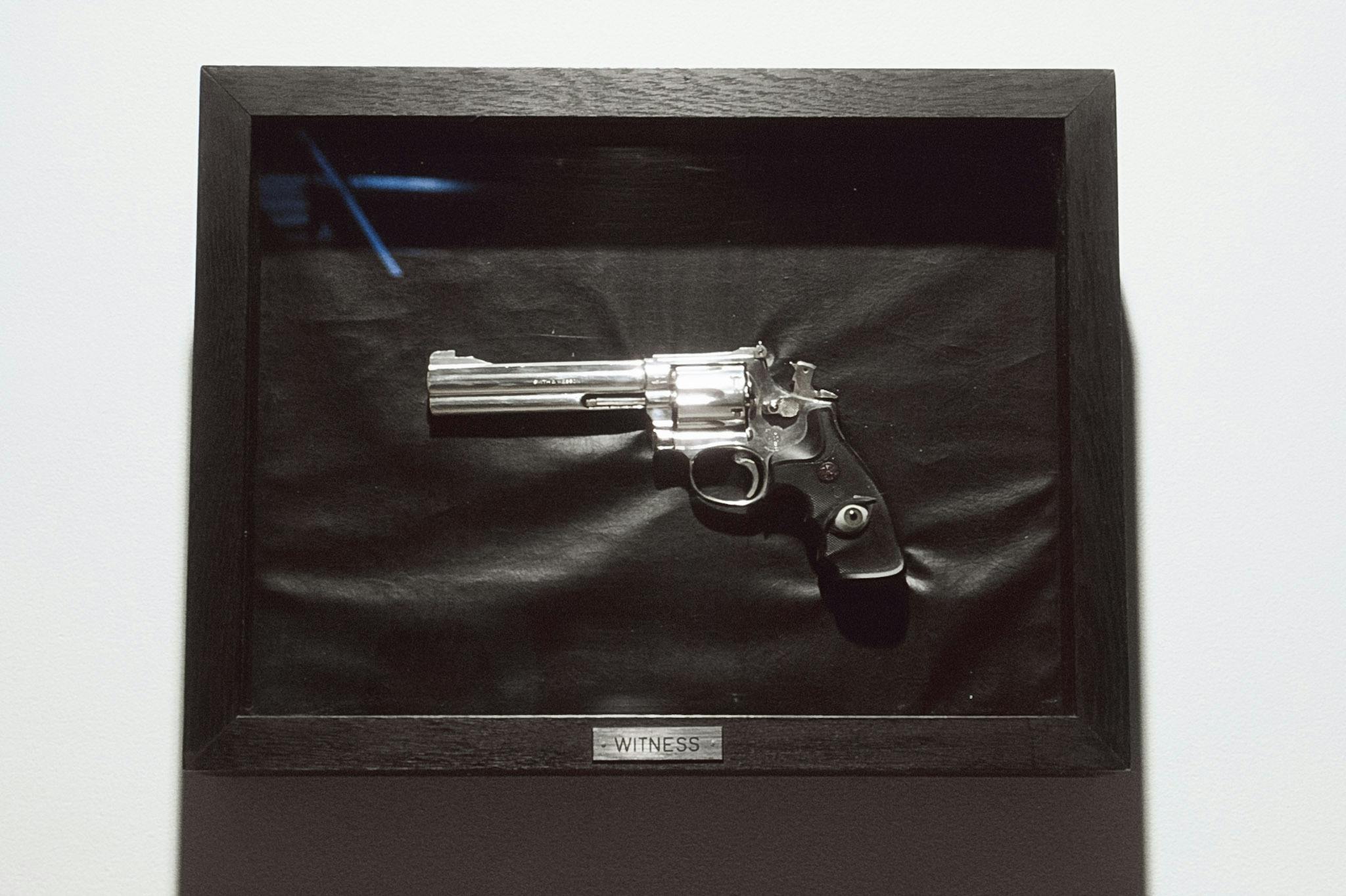 A silver revolver gun is framed in black and installed on a gallery wall. The word, WITNESS is carved on a small silver plaque nailed to the frame. A fake human eyeball is inlaid in the grip. 