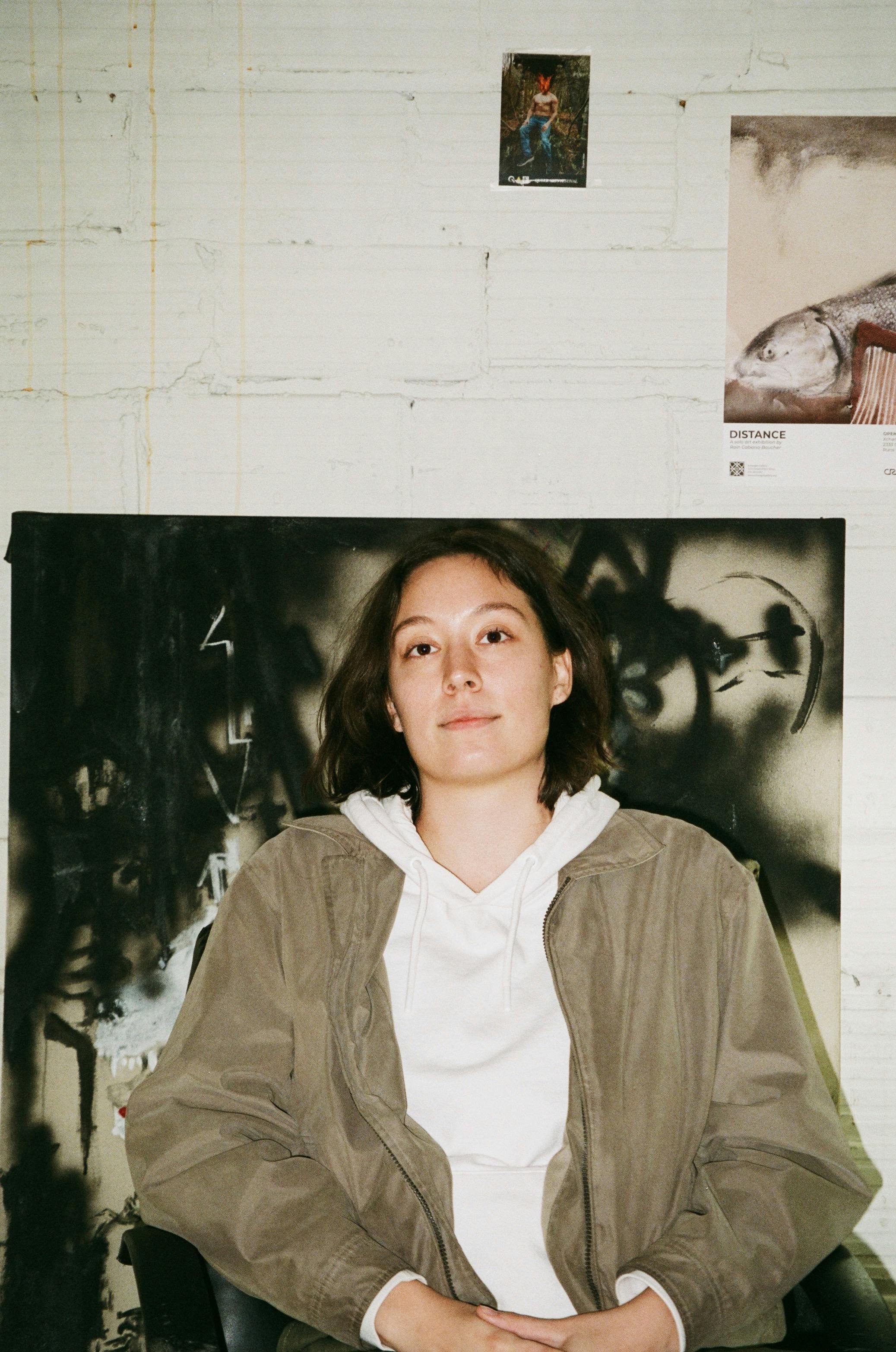 A portrait of Rain Cabana-Boucher. She is seated in front of a black and white painting hung on a white brick wall, on which two smaller images hang.