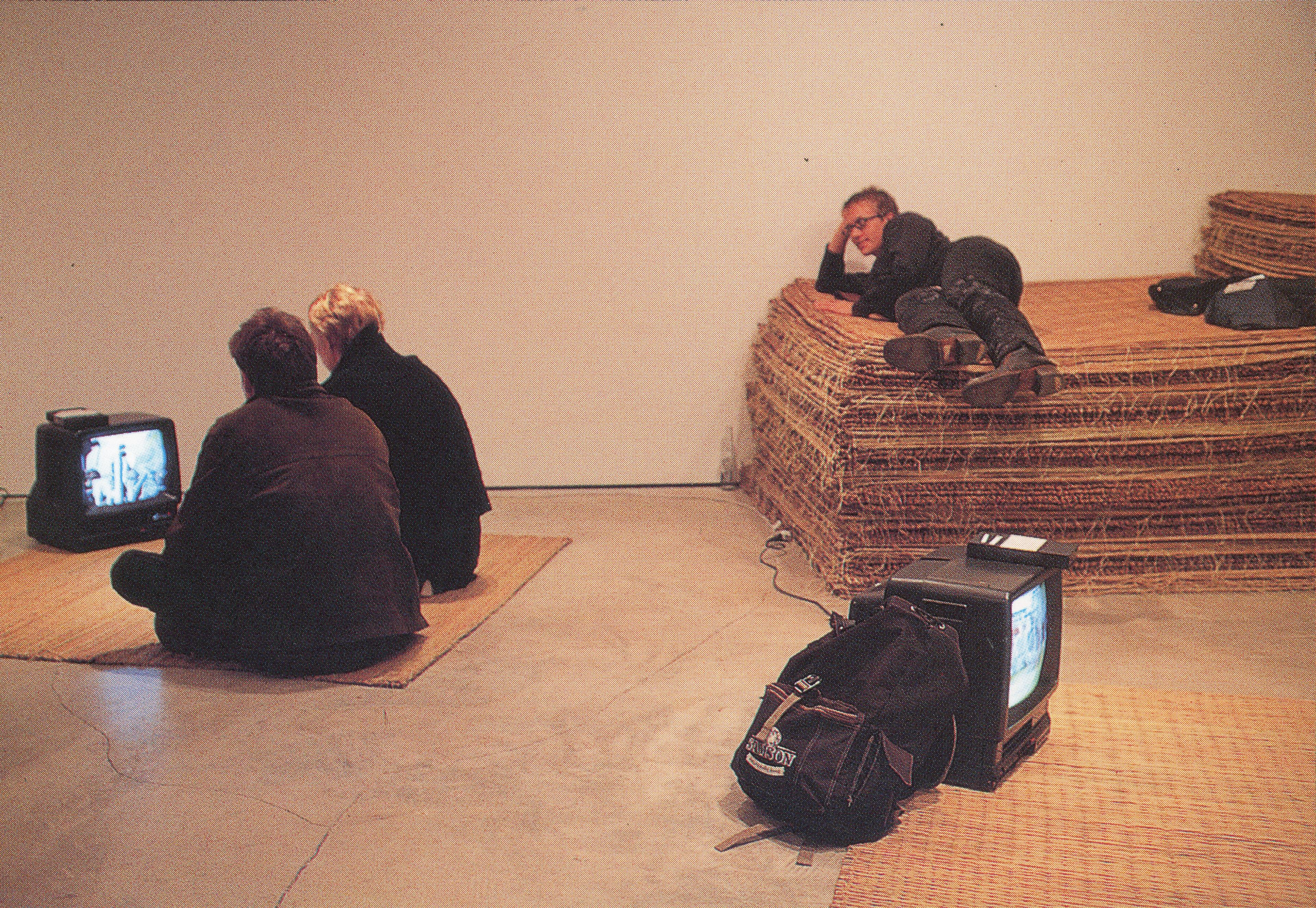 A rush mat and a CRT TV are placed on a gallery floor. Two people sit on a mat and are watching the video played on the TV. Behind them, a person lies on a pile of rush mats.