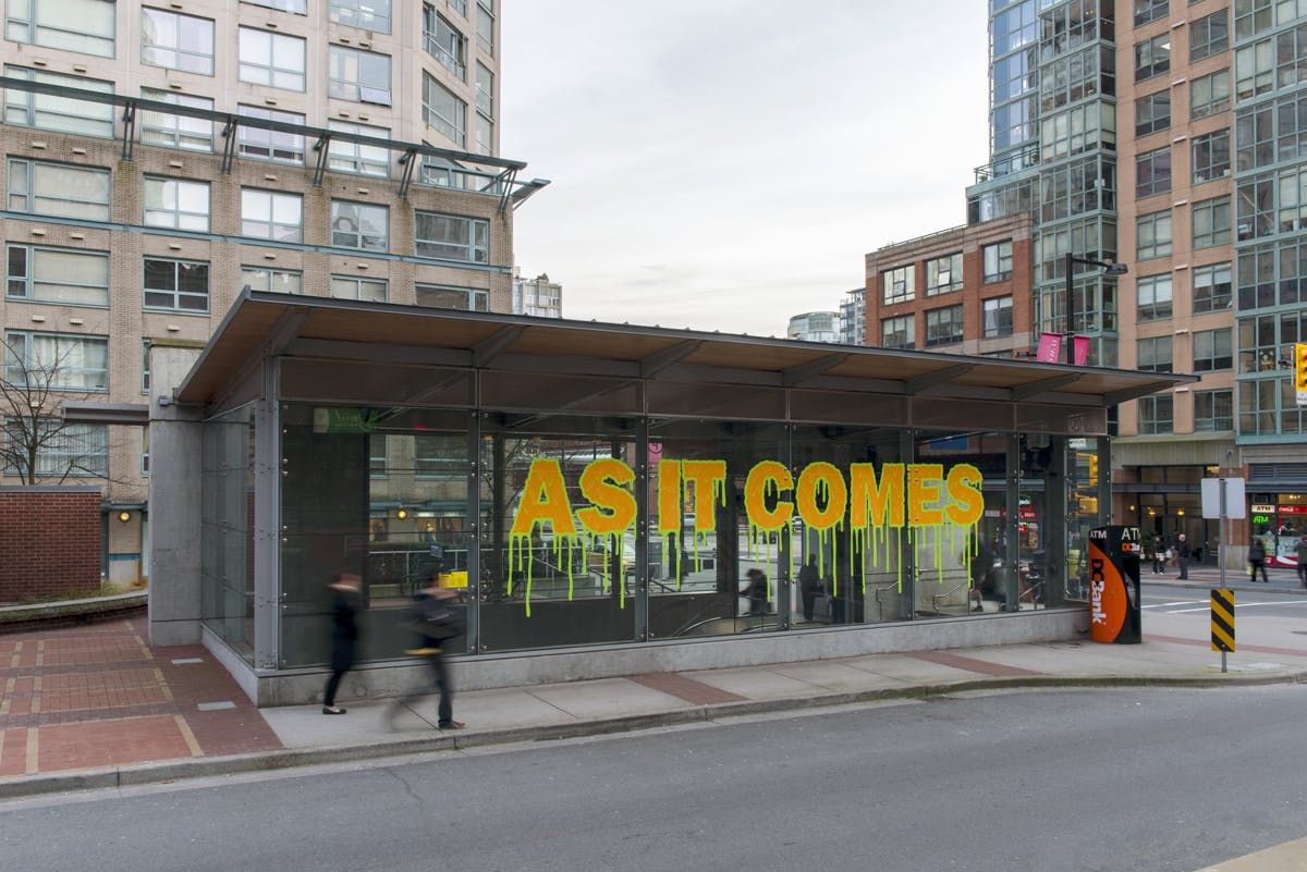 Text in vinyl installed on the glass exterior of Yaletown-Roundhouse Station. “AS IT COMES” appears in orange-yellow colour. Letters are outlined by dripping, neon green lines.