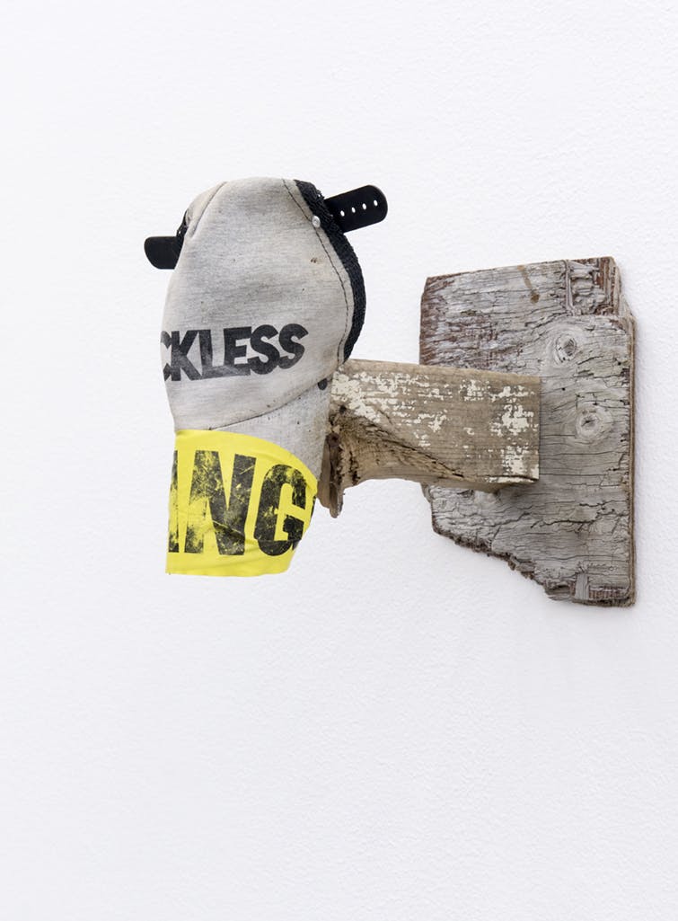 A grey cap hangs from a short wooden beam installed vertically on a gallery wall. The cap has a black plastic snap which is modified to make the cap look like a cow’s face.