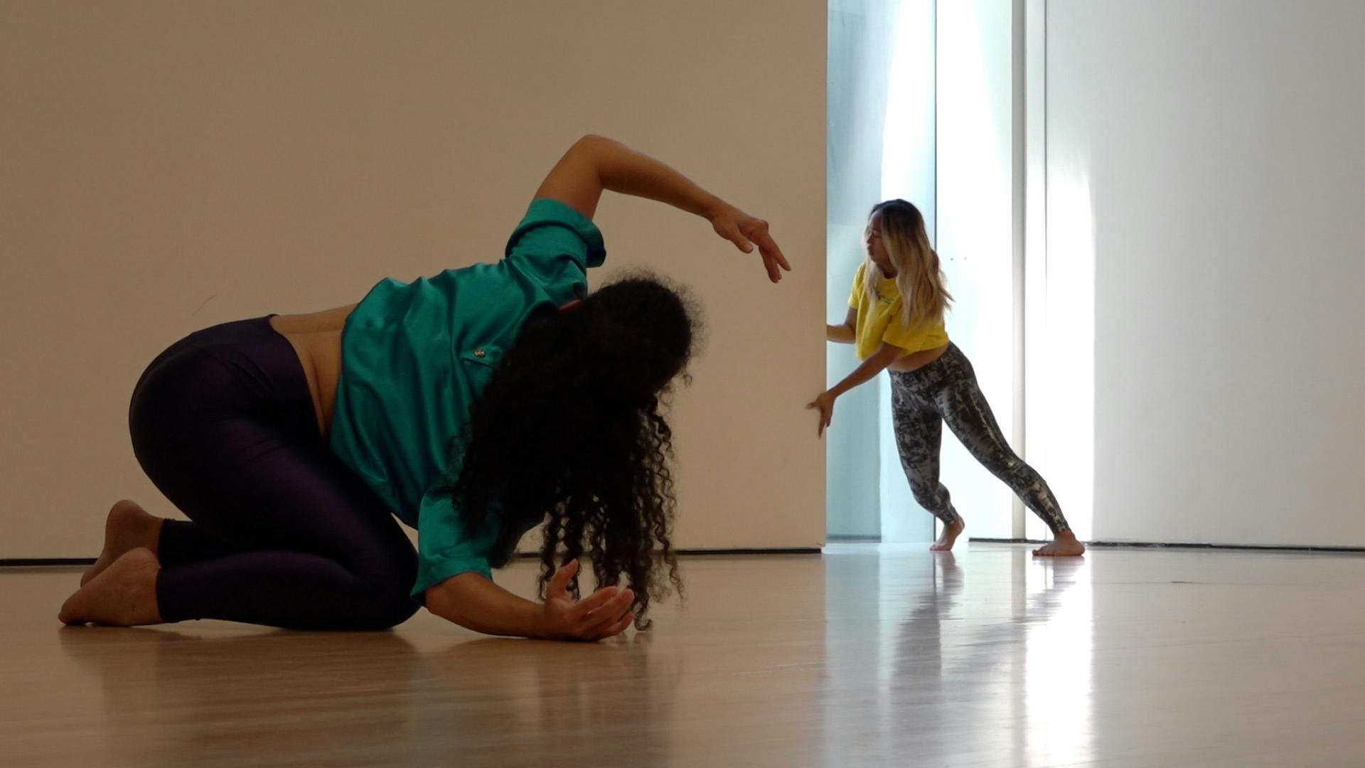 A video still of two dancers performing in a studio space. In the foreground, a dancer is crouched on their knees with one arm sweeping overhead. In the background, a dancer is holding the corner of a wall, leaning forward with their feet planted.