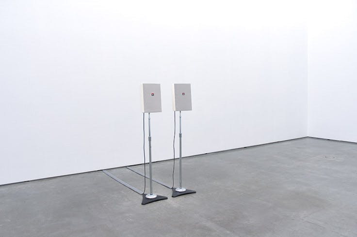 An installation view of a work titled Stereo by Ceal Floyer. Two identical speakers on tall stands are placed in the middle of a gallery space.