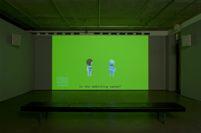A single-channel video is projected on a wall in a darkened gallery space. The scene depicts naked, toy-like figures speaking to each other. The subtitle reads, “In the addicting sense?”
