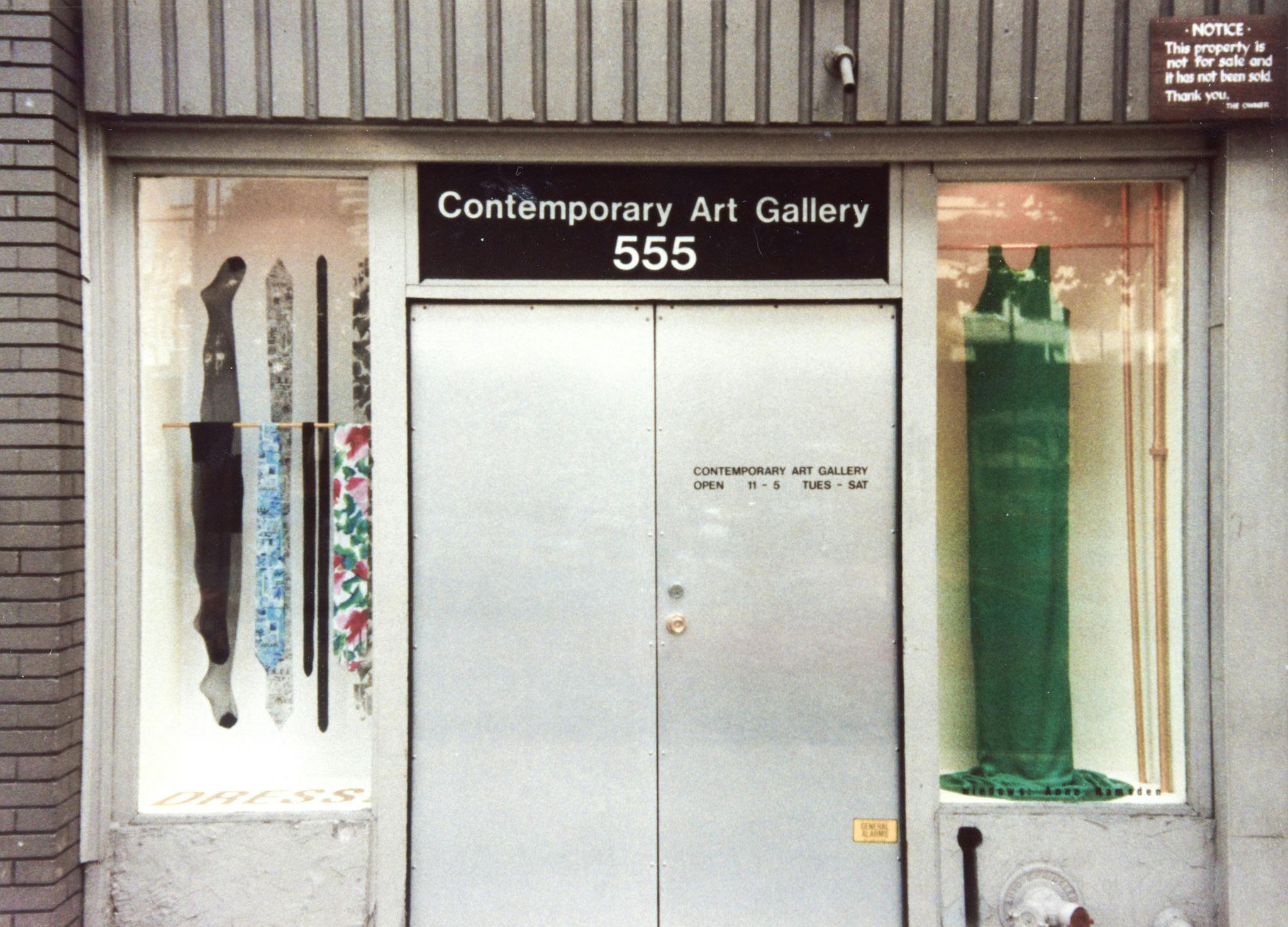 The facade of a gallery, with artworks in the window display. The works in the windows consist of garments hanging on metal rods, including pantyhose, neckties, a belt, and a bright green dress. 