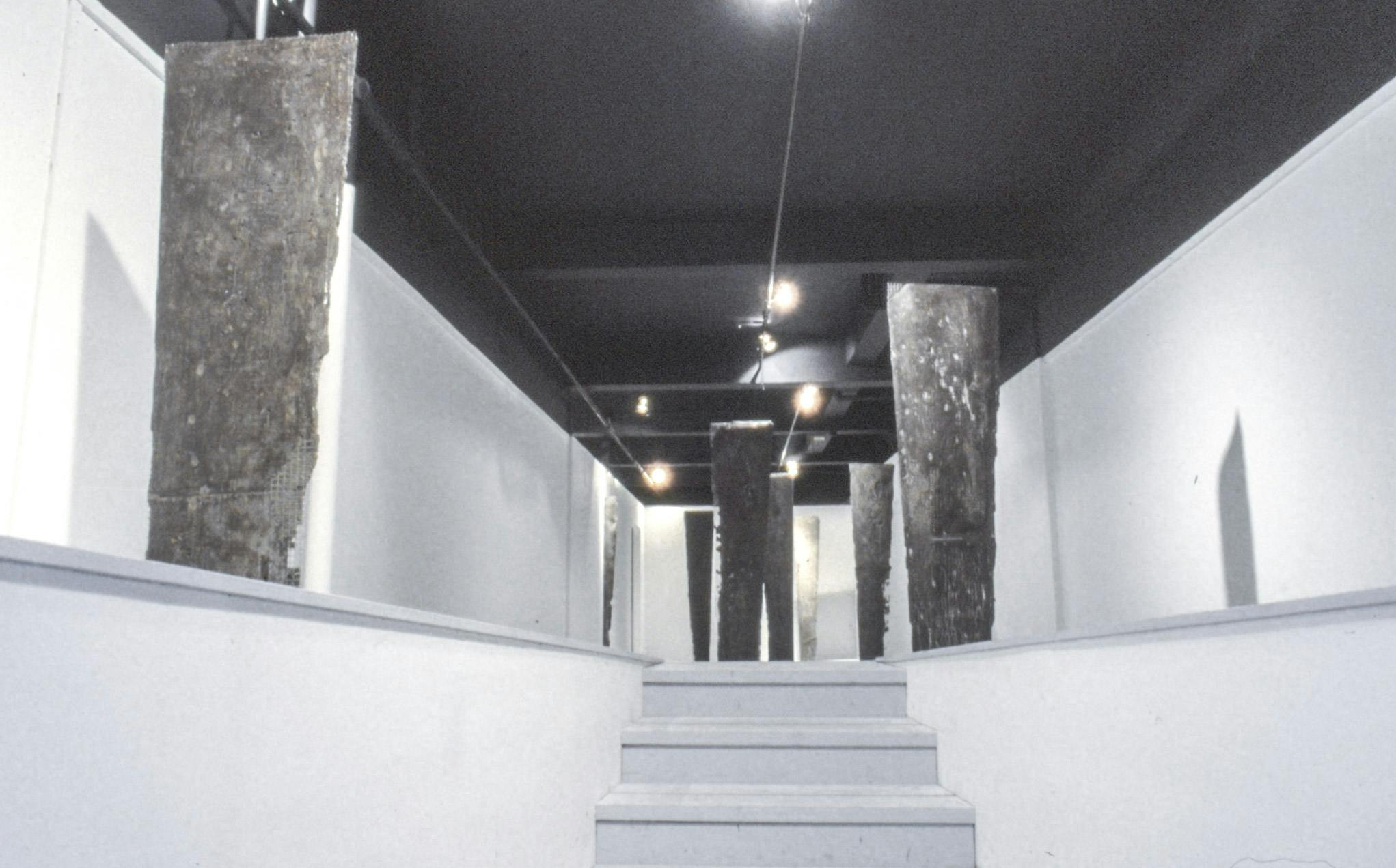 In a gallery space with a dark ceiling, there is a small set of steps leading to a white platform. On the platform, there are several planks of metal-cement with one straight edge and one jagged edge.