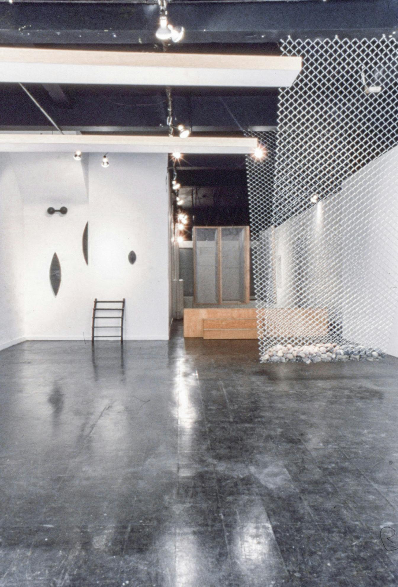 Artworks in a gallery. In the foreground, there is a suspended metal net with rocks. There are small black objects mounted on one wall, and a wooden platform with a closet-like form on top of it.