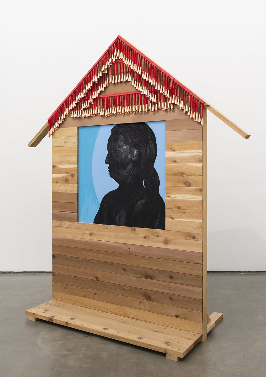 A large wood sculpture is installed in a gallery space. It is the shape of a house with a large window in the middle. A painting depicting the silhouette of a woman sits in the window.