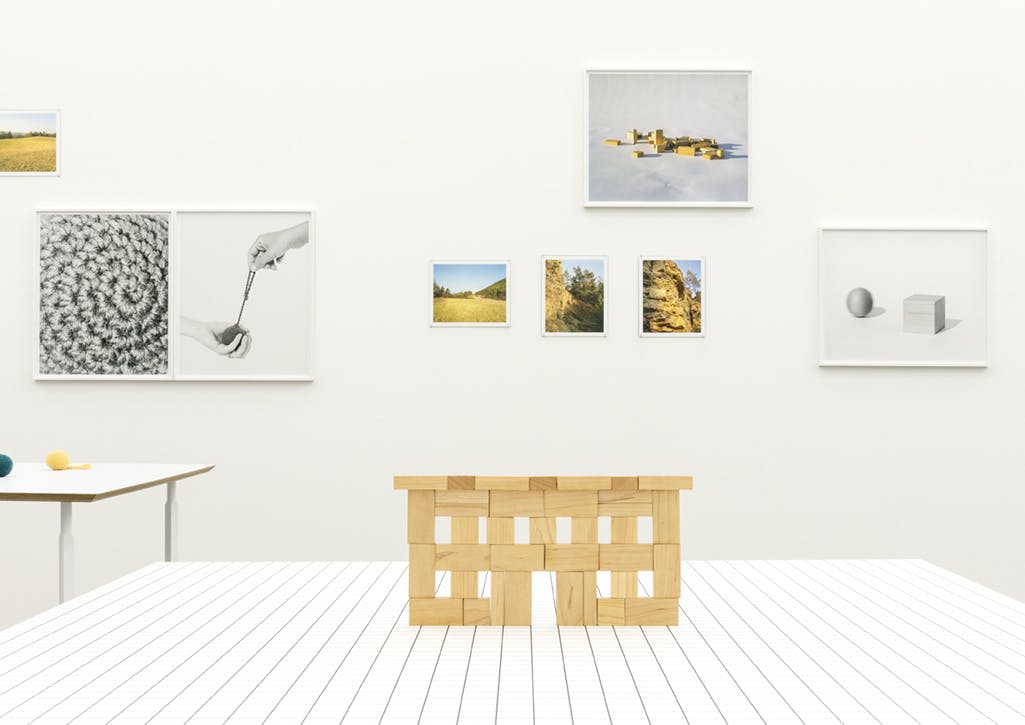 Stacked wooden blocks on a white table resemble a simple facade structure. Several framed photographs of landscapes, wooden objects and natural materials hang on the wall behind.