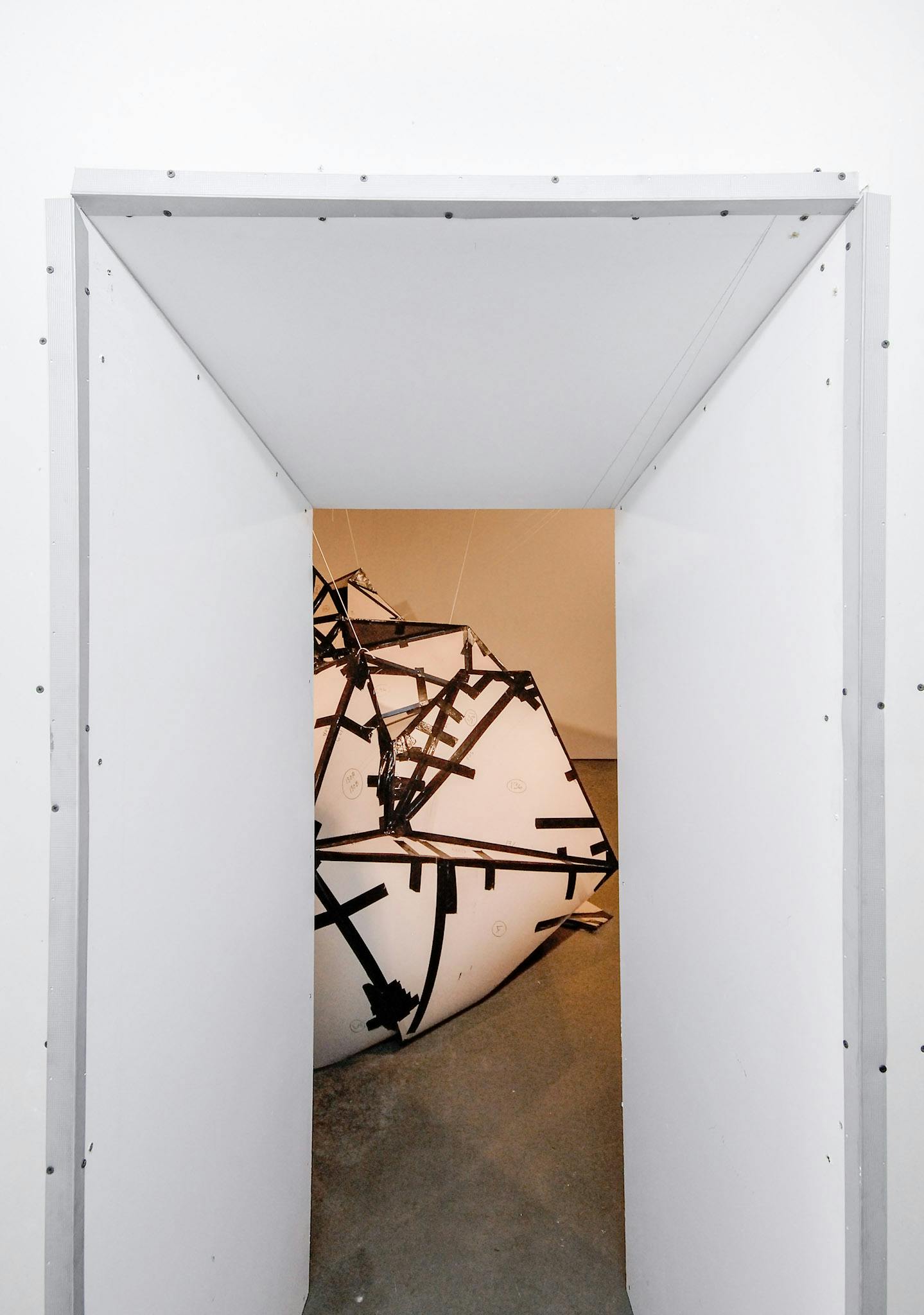 This is an entrance to a gallery space, which is narrowed by white panels attached above the gallery entrance. A large-sized white sculpture is partially visible through the entrance. 