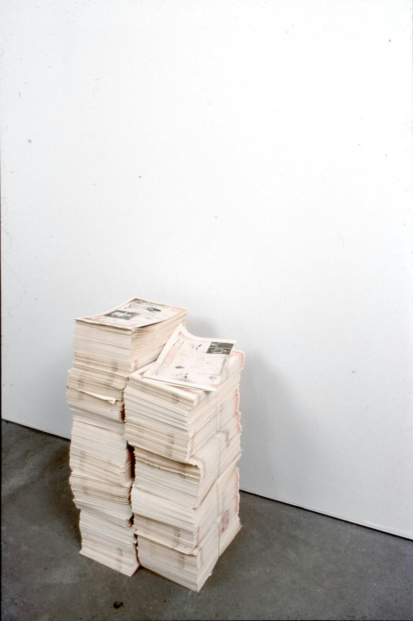 Two piles of newspaper-style publications are placed directly on a gallery wall. They are the publication titled I Got Killed, I Got Killed, I Got Killed in World War III that accompanied this group exhibition.