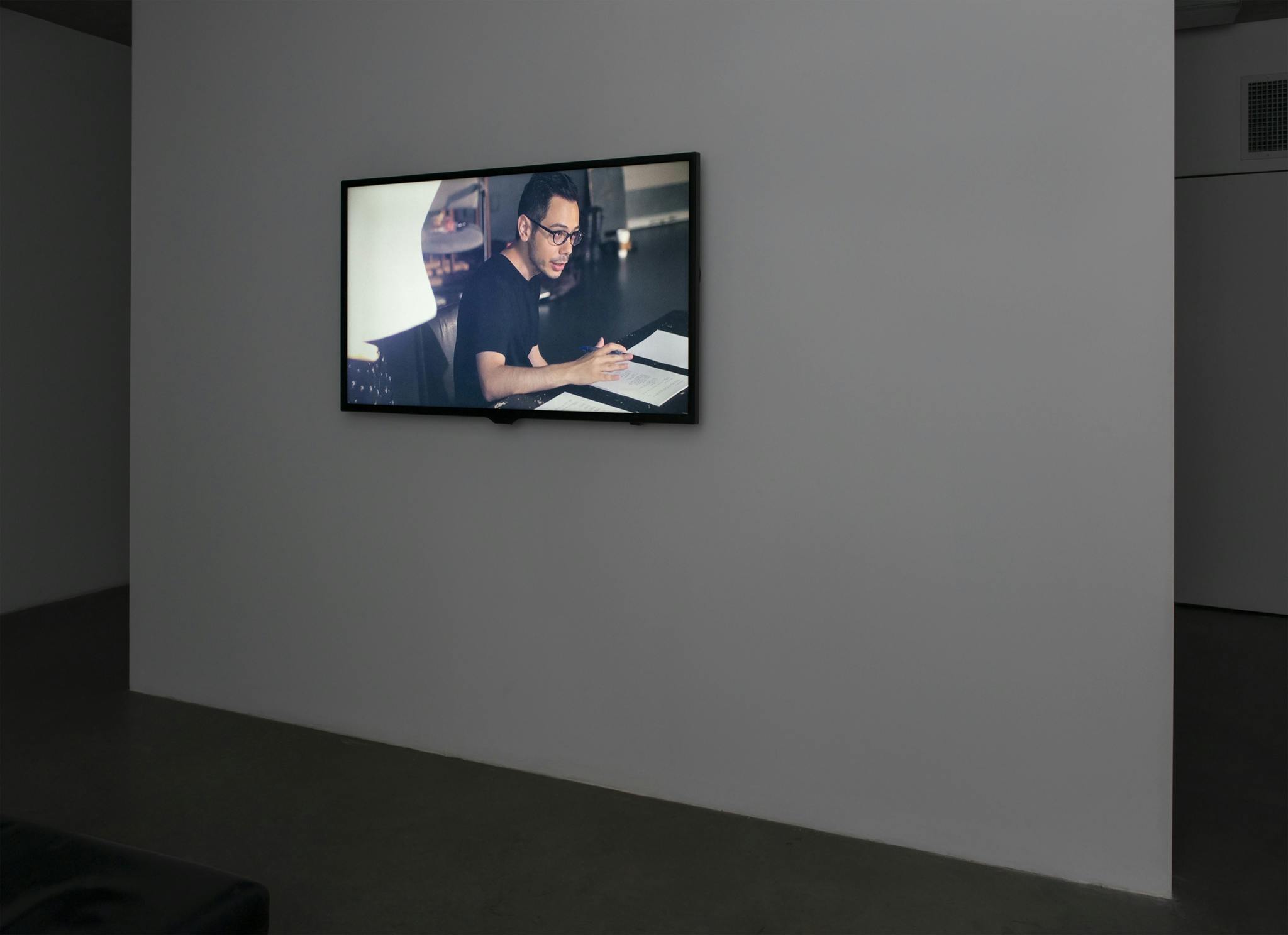 A monitor displaying a video work hangs on the wall of a dark gallery space. Onscreen, an actor sits at a table, auditioning for an imagined documentary.