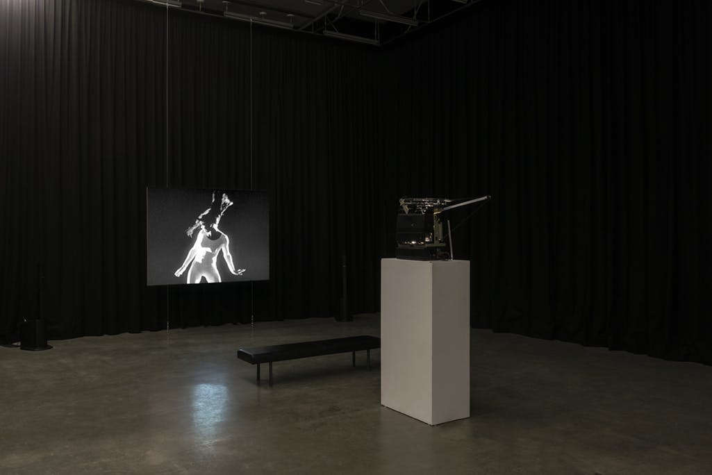 In a gallery enclosed by black curtains, a single-channel video is projected onto a screen. The projector is atop a plinth in front of the screen. In the video, a person in a white leotard dances.
