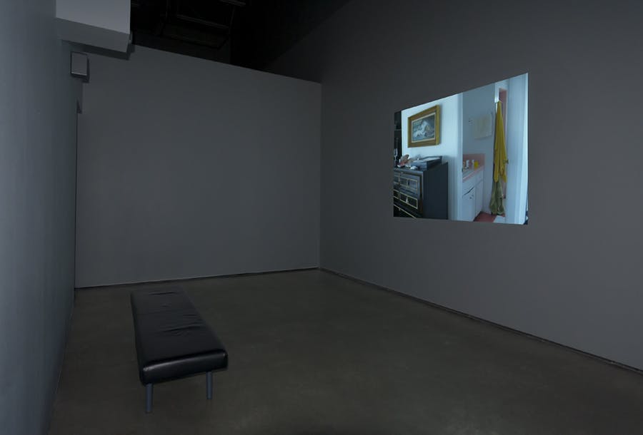 Installation image of a projected image onto a wall in a dimly lit gallery. A black bench is placed in front. The image depicts a corner view of a bedroom’s dresser and en suite bathroom vanity. 