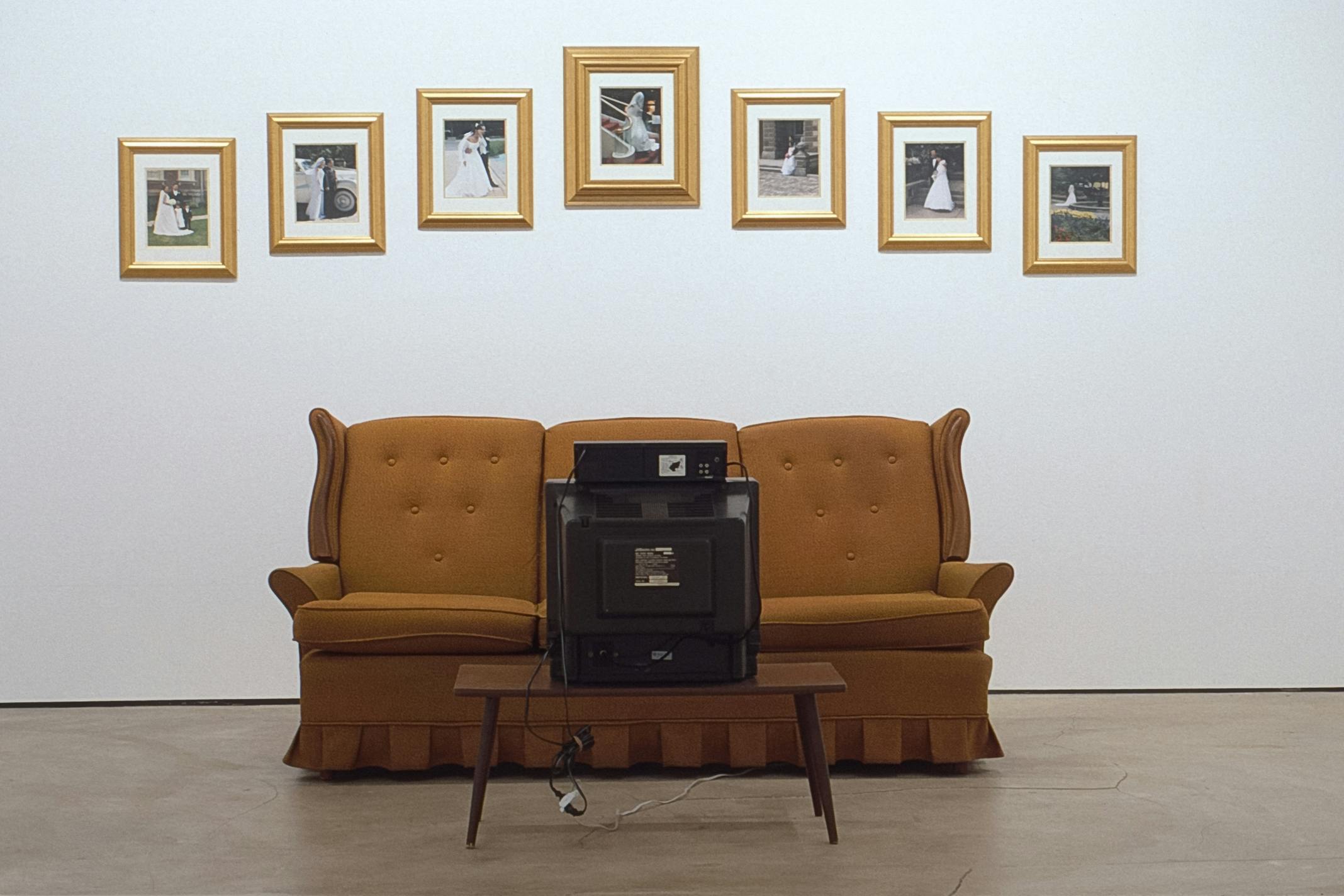Seven wedding photographs are installed on the gallery wall above the mustard-coloured couch. A black CRT TV is placed in front of the couch. All photographs are gold framed. 