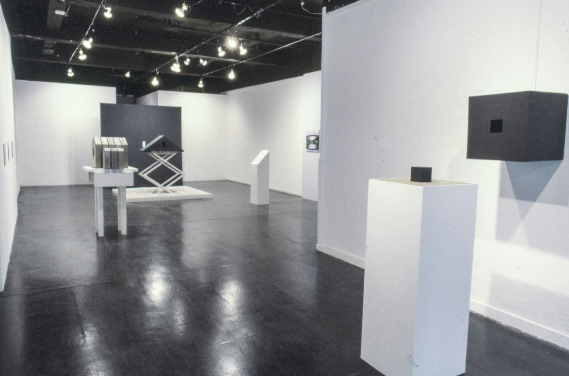 Several sculptures and small paintings in a gallery space. The sculptures are black and concrete, and are on several different white surfaces including a plinth, a side table, and a scissor lift.