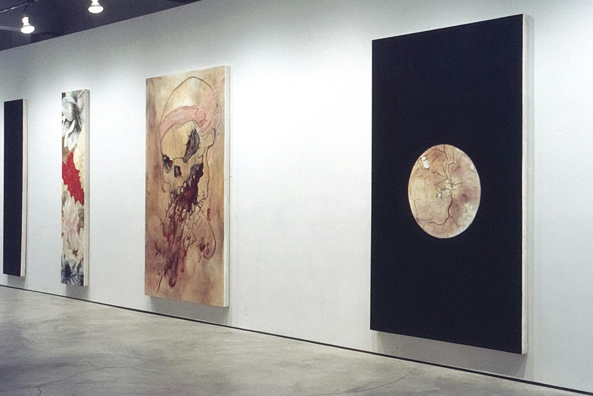 Four vertical paintings in a gallery. One is all black, one has several large flowers layered together, one has a skull spewing blood from its mouth, and one is black with a textured beige circle. 