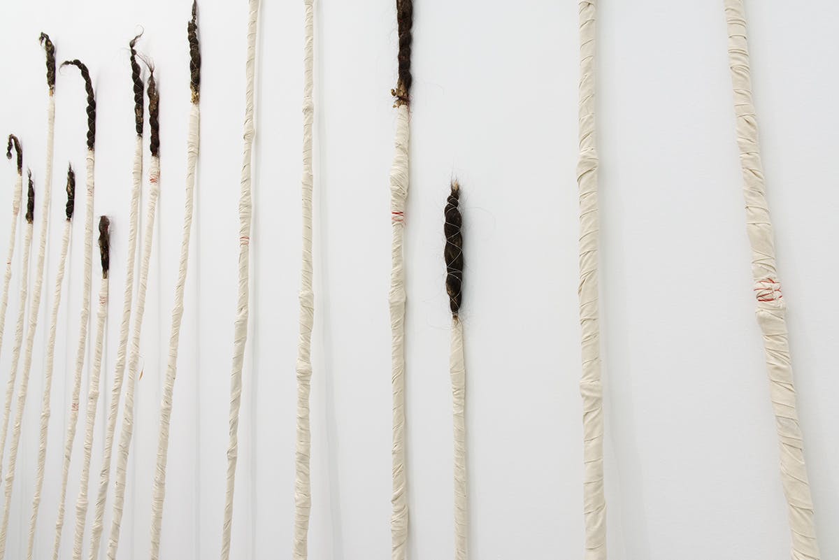 This is the side view of a set of Charlene Vickers’ wrapped sculptures leaning against a wall. The length of each sculpture varies. Human hair emerges from the top of each sculpture.