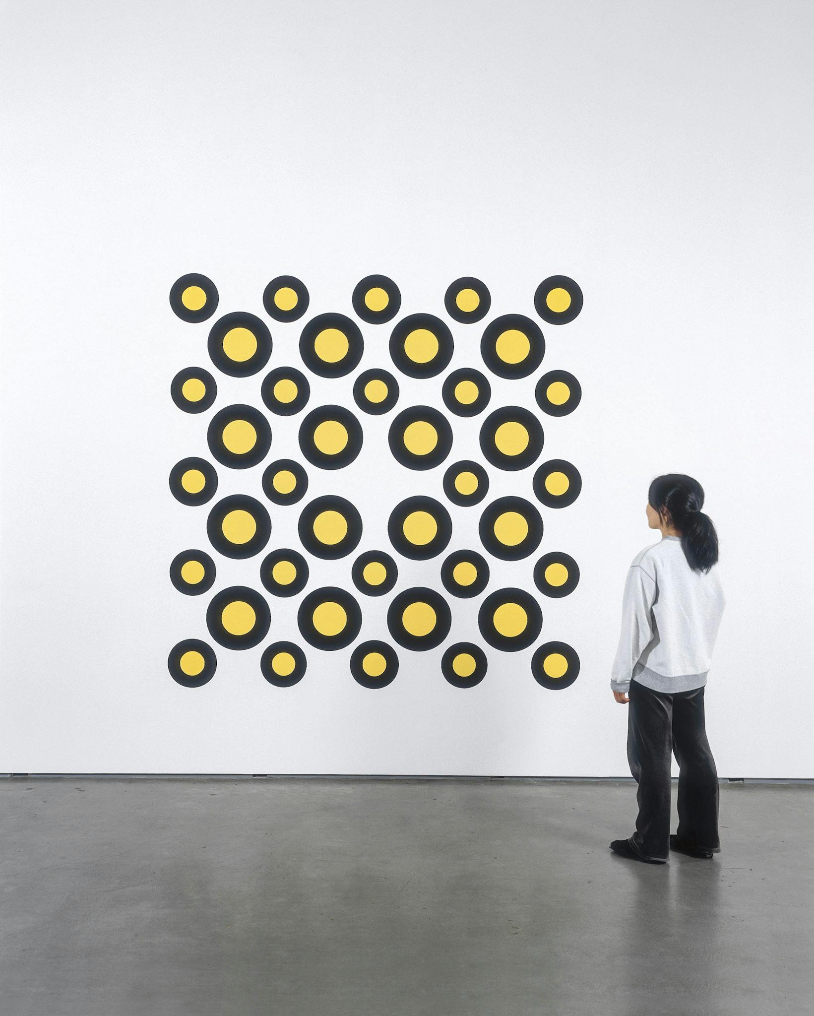 Installation image of a painting by Neil Campbell. The work consists of many yellow circles outlined by thick bold black lines. The painting is as tall as a person standing near it.