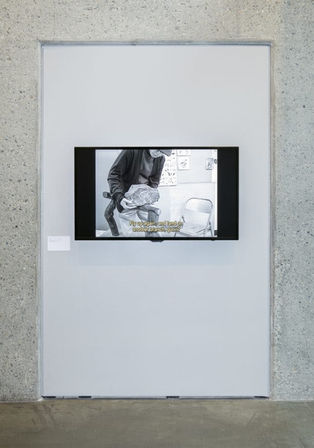 Detail view of a gallery wall framed by light gray concrete. In the middle, a monitor displays a black and white video, depicting an older person trying to lift or carry a large rock.