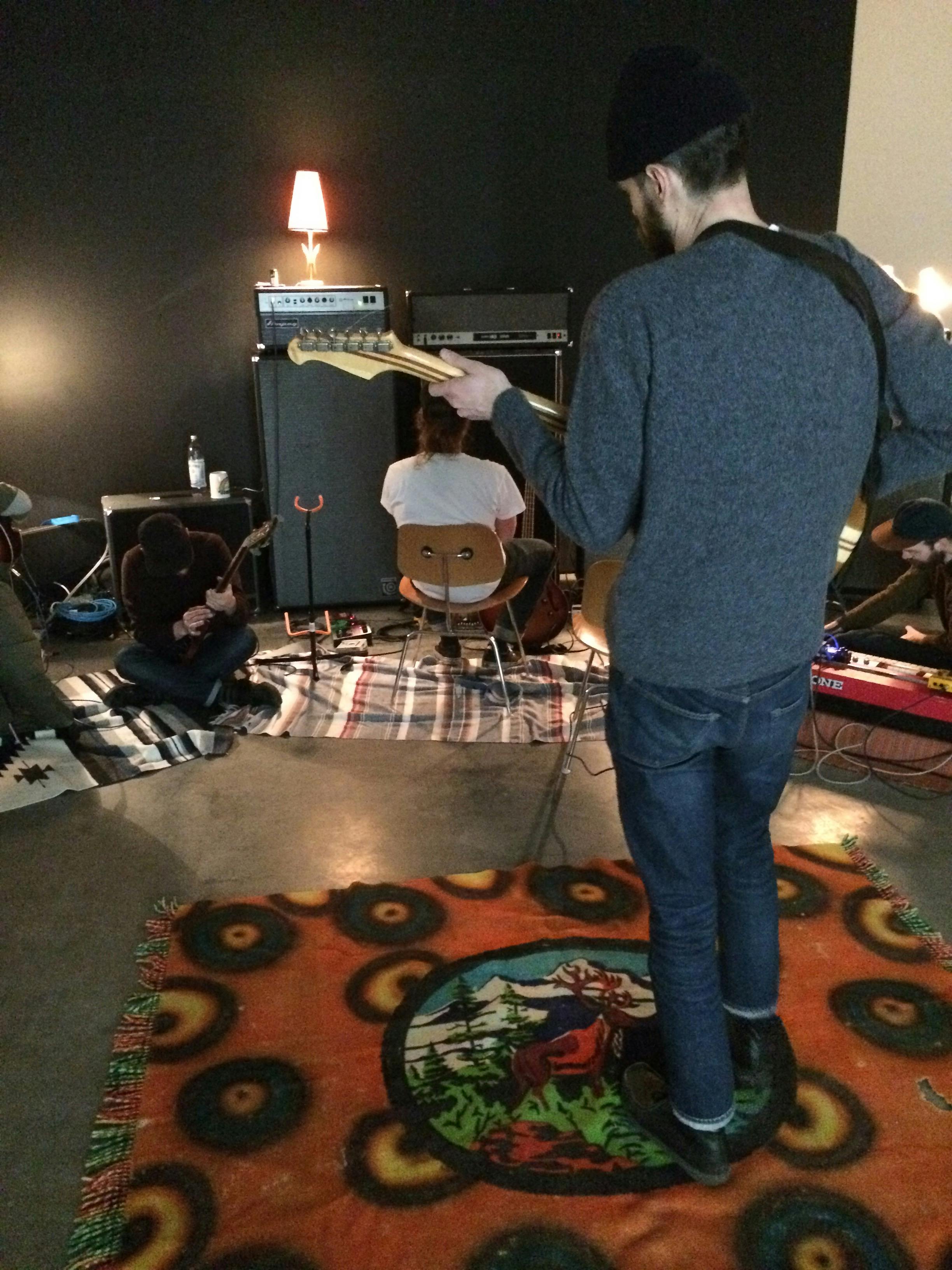 An image of four people setting up their recording studio. Some people hold guitars. Each musician sits or stands on a blanket.