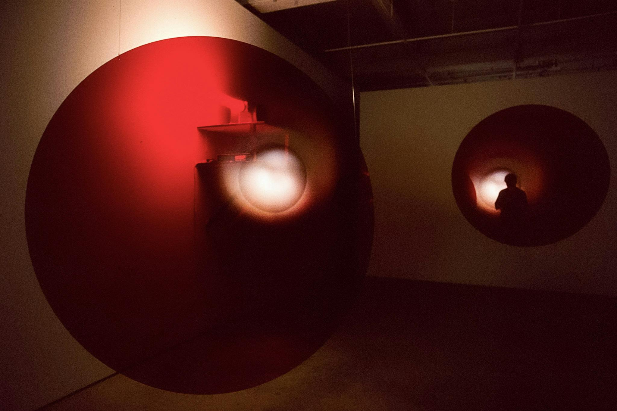 Two large circular-shaped thin red sculptures are visible in this photograph. Hanging from the gallery’s ceiling, these sculptures have a weak white light source placed slightly off-centered. 